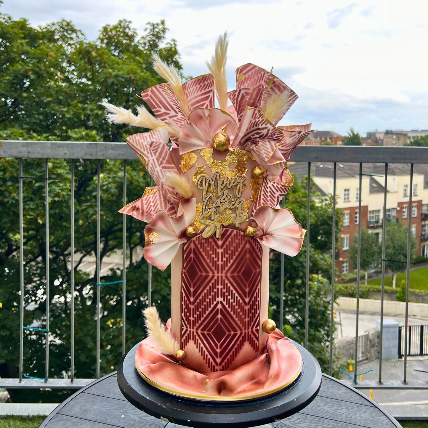 Decorative cake with beige and gold pattern on wafer paper on a balcony with greenery and buildings in the background