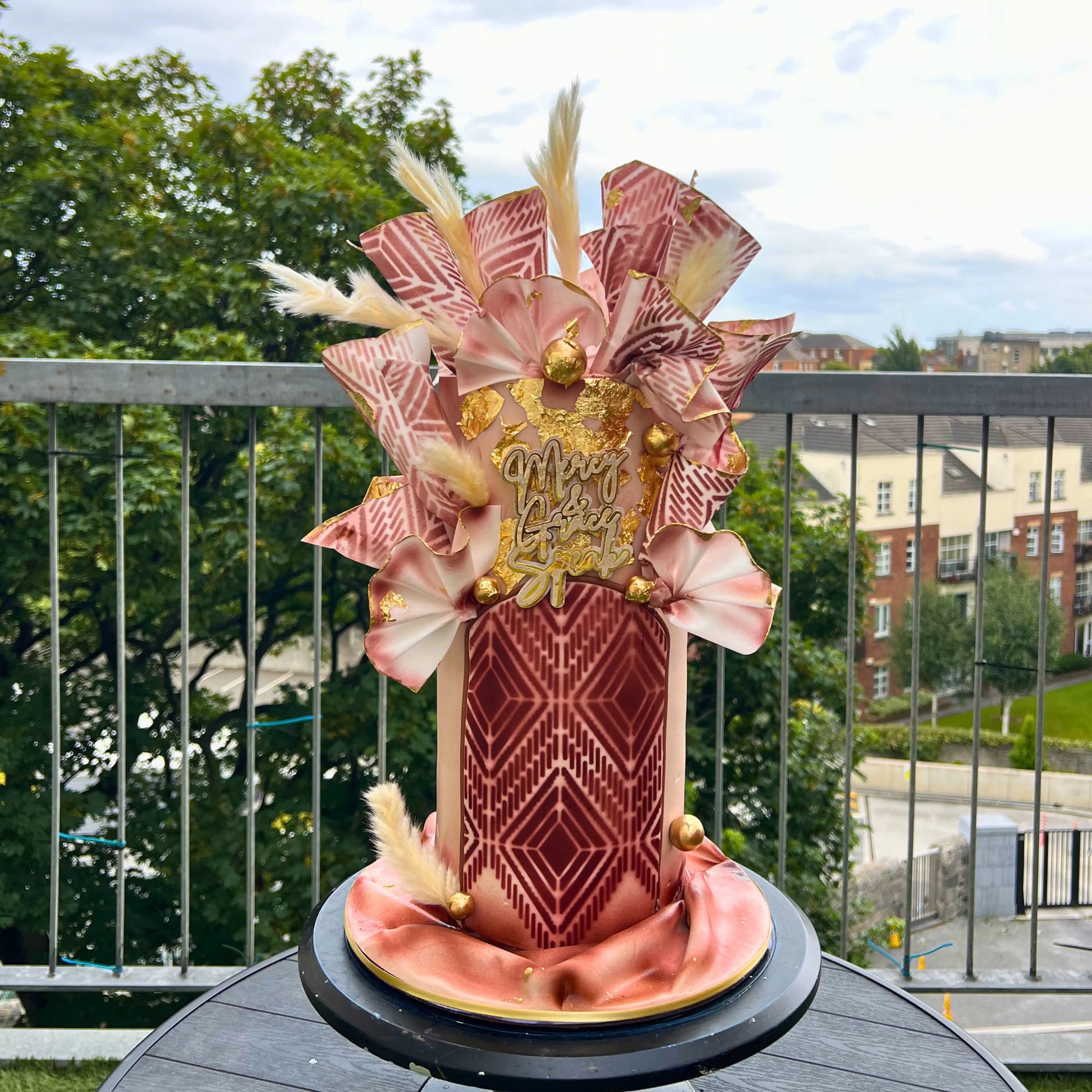 Decorative cake with beige and gold pattern on wafer paper on a balcony with greenery and buildings in the background