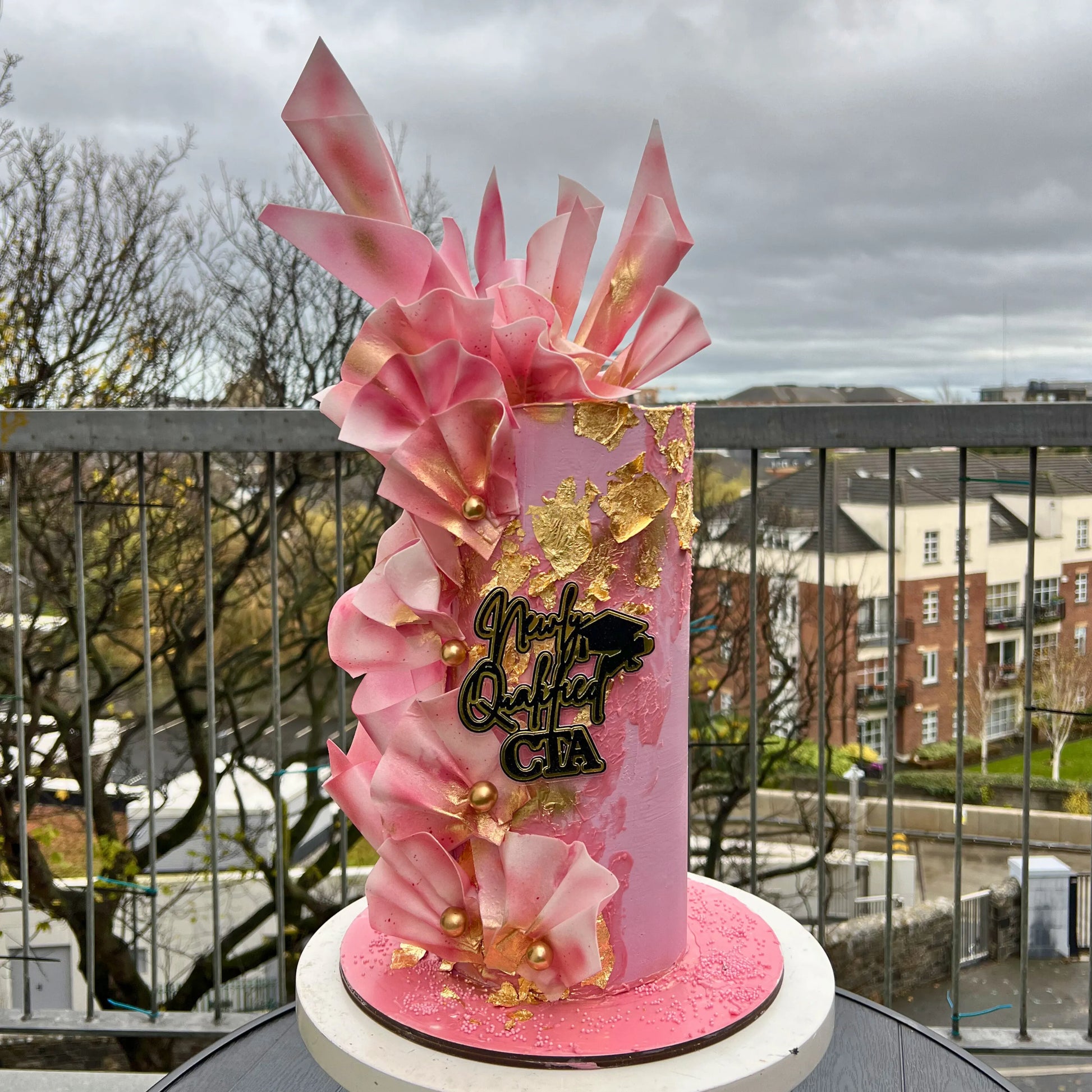 Decorative pink and gold'Nikki graduation cake on a balcony with a cityscape background.