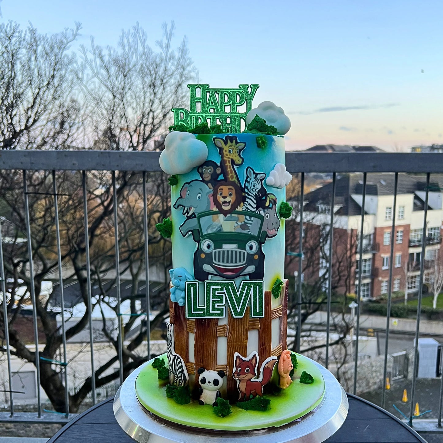 Children's birthday cake with animal-themed decorations and 'Happy Birthday Levi' topper on a balcony.