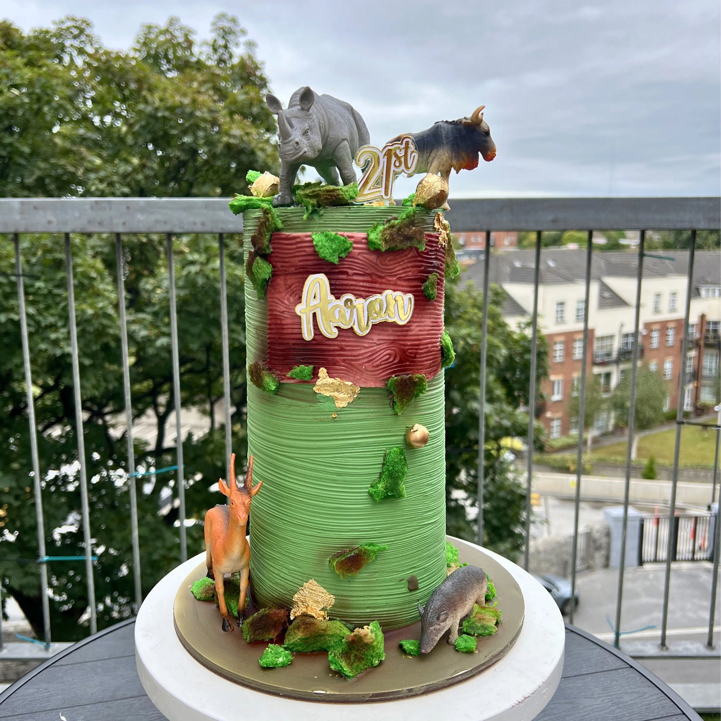 Decorative cake with animal figures and '21st' on a balcony with trees and buildings in the background