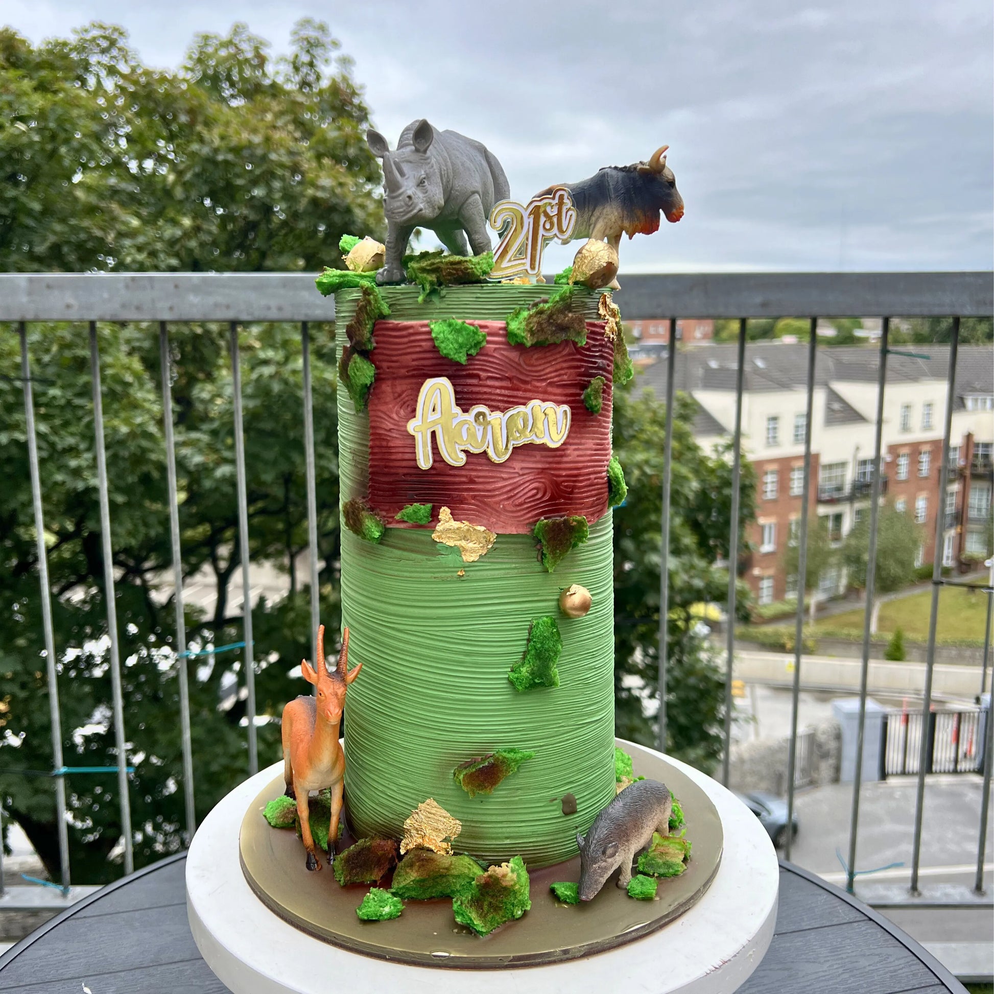 Decorative cake with animal figures and '21st' on a balcony with trees and buildings in the background
