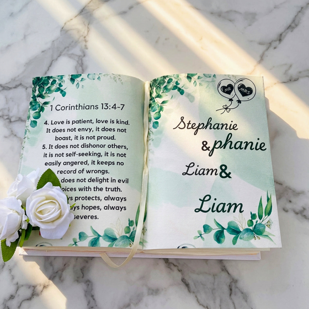 An open personalized Bible journal on a white marble surface, featuring printed scripture on one page and a custom name page on the other with watercolor green leaves and white roses as props.