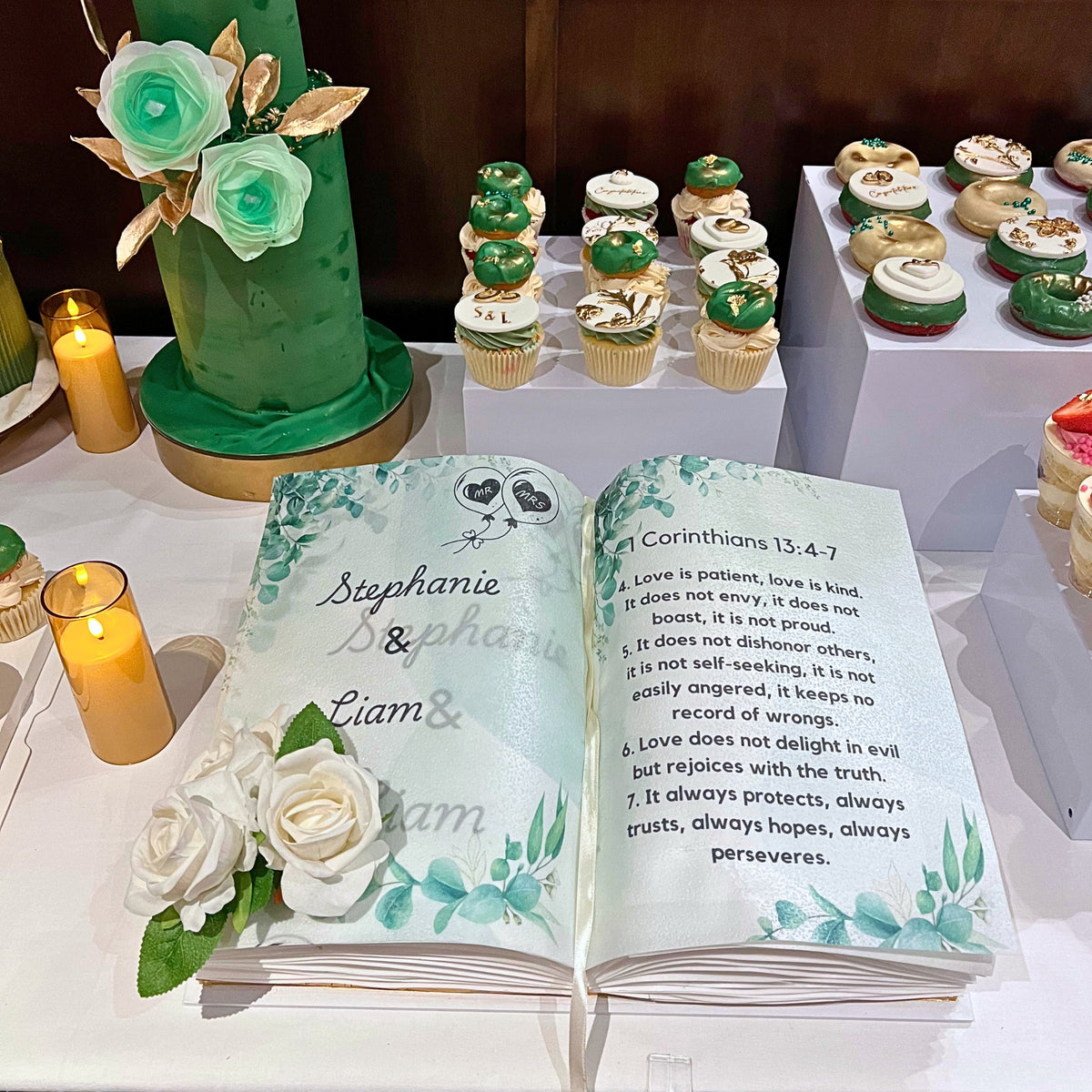 A custom wedding book cake, styled on a white tablecloth, surrounded by green and white cupcakes, candles, and a green cake. The open guest book features floral watercolor designs and Bible verse text.