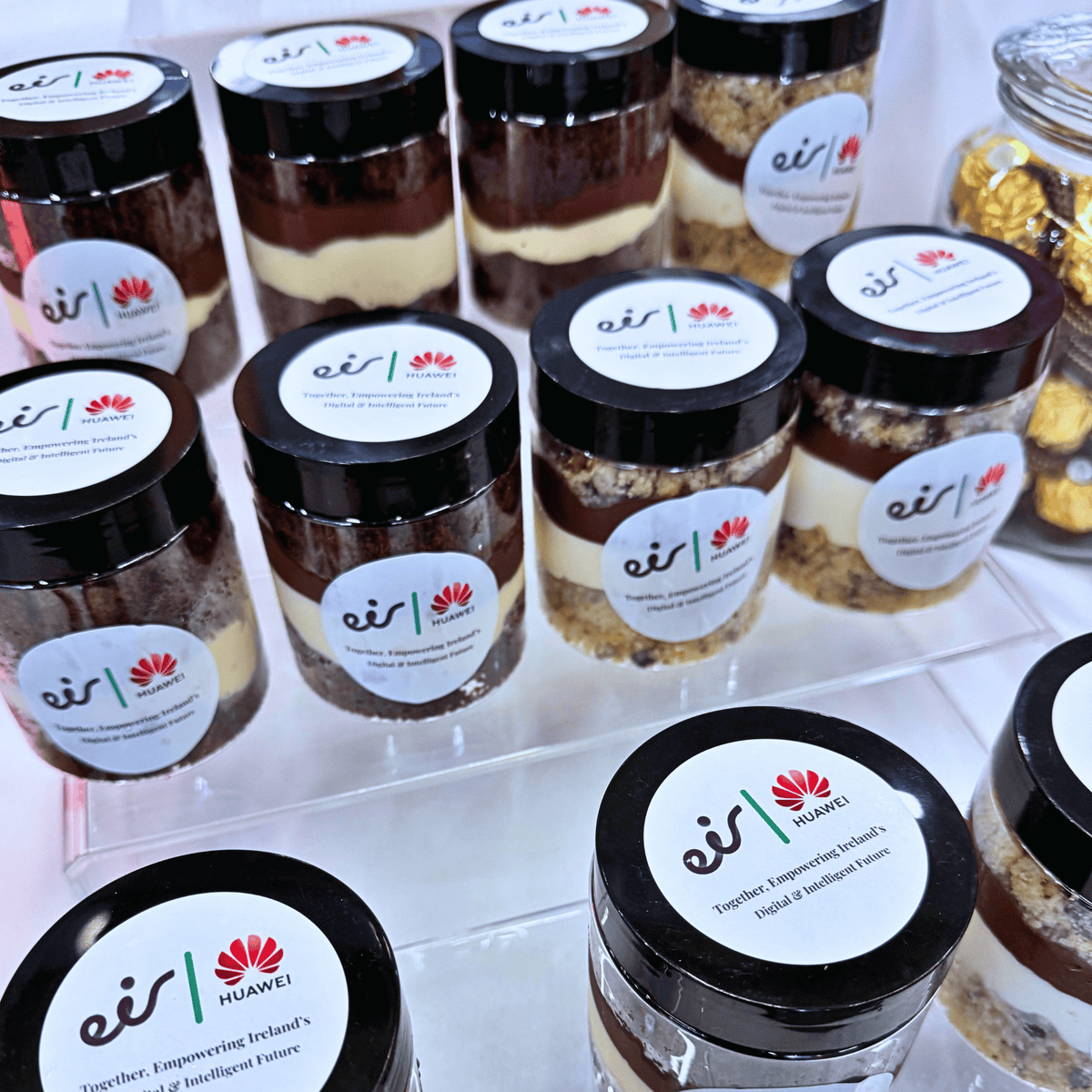 Several glass jars of layered dessert for a corporate event in Dublin city. Each jar has a black lid and a white label featuring the 'eis | HUAWEI' logo and tagline 'Together Empowering Ireland's Original Intelligent Future'.