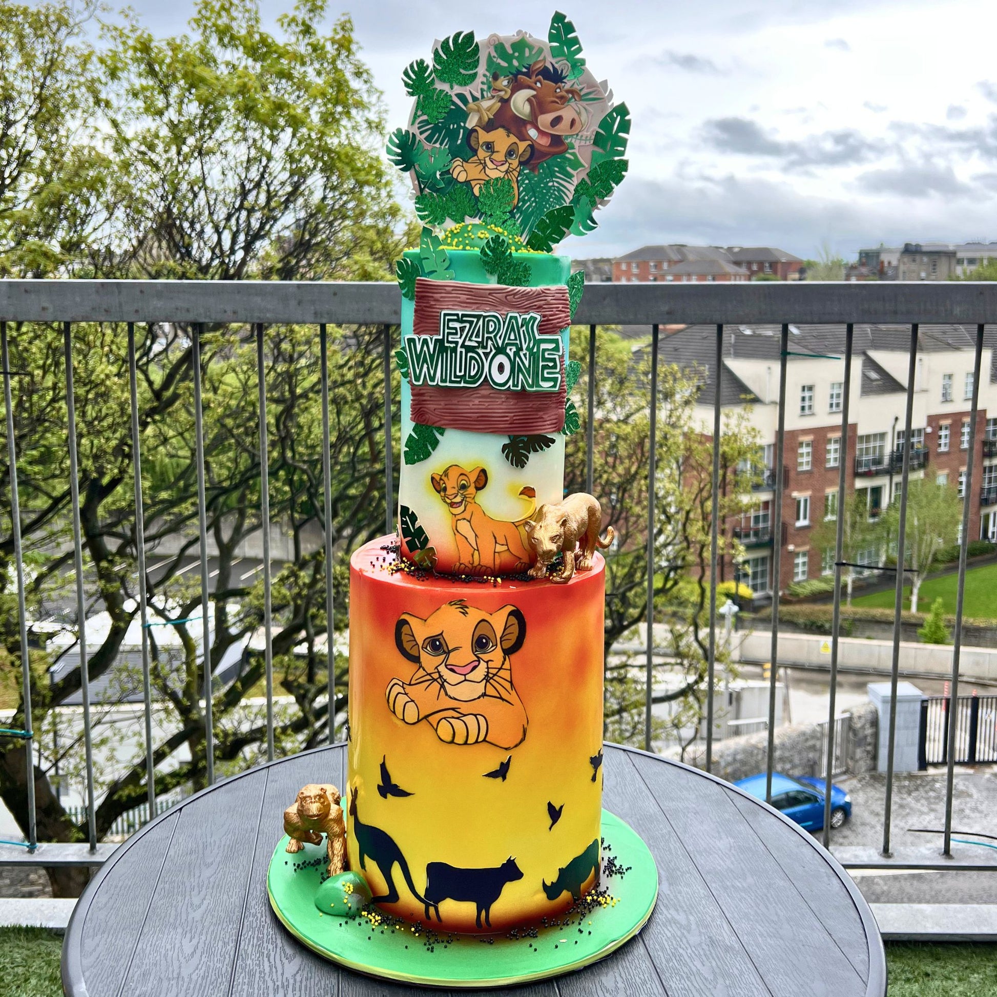 A custom two-tiered cake decorated with Simba and Nala from The Lion King, displayed on a balcony table with a city background visible through the railing.
