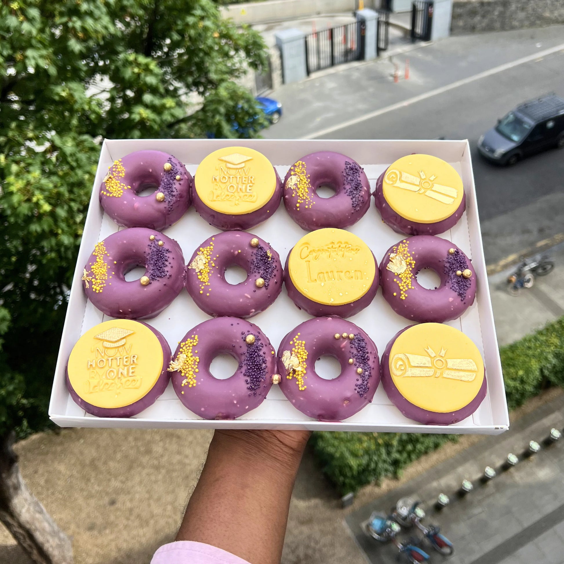 Box of purple and yellow donuts held by a person outdoors.