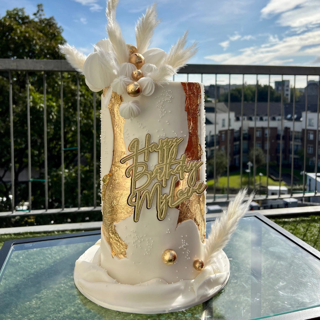 A decorated cake with white and gold fondant and edible wafer paper sails, with a 'Happy Birthday' message in gold lettering, placed on a glass table with a balcony view in the background.