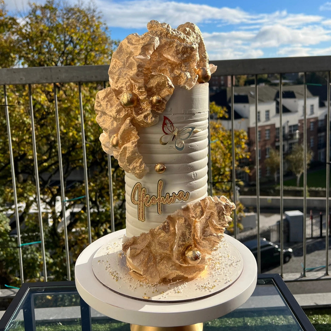 A decorated buttercream cake with gold wafer-like icing, presented on a glass cake stand .