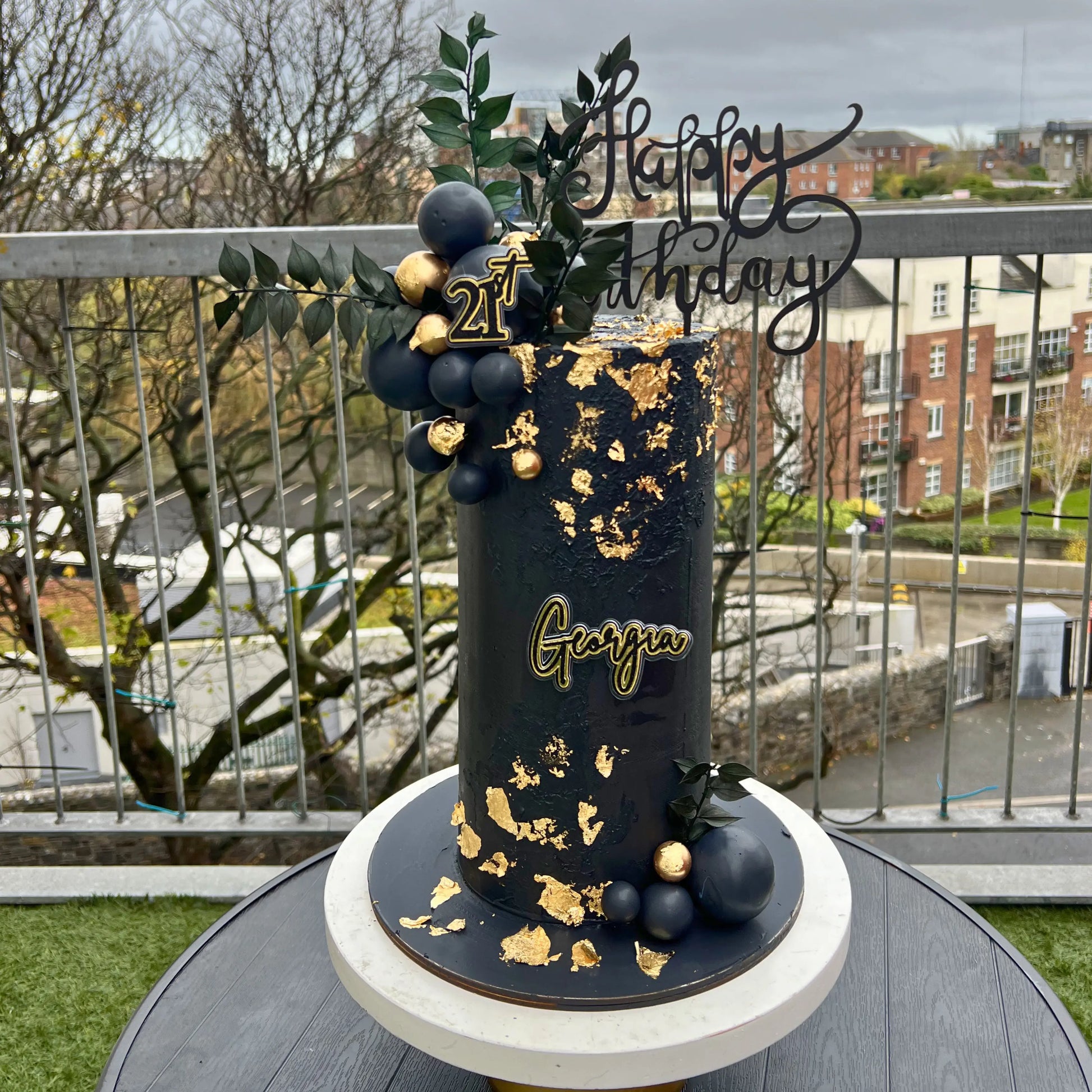 Decorative cake with gold and black design on a balcony with a view of buildings and trees.