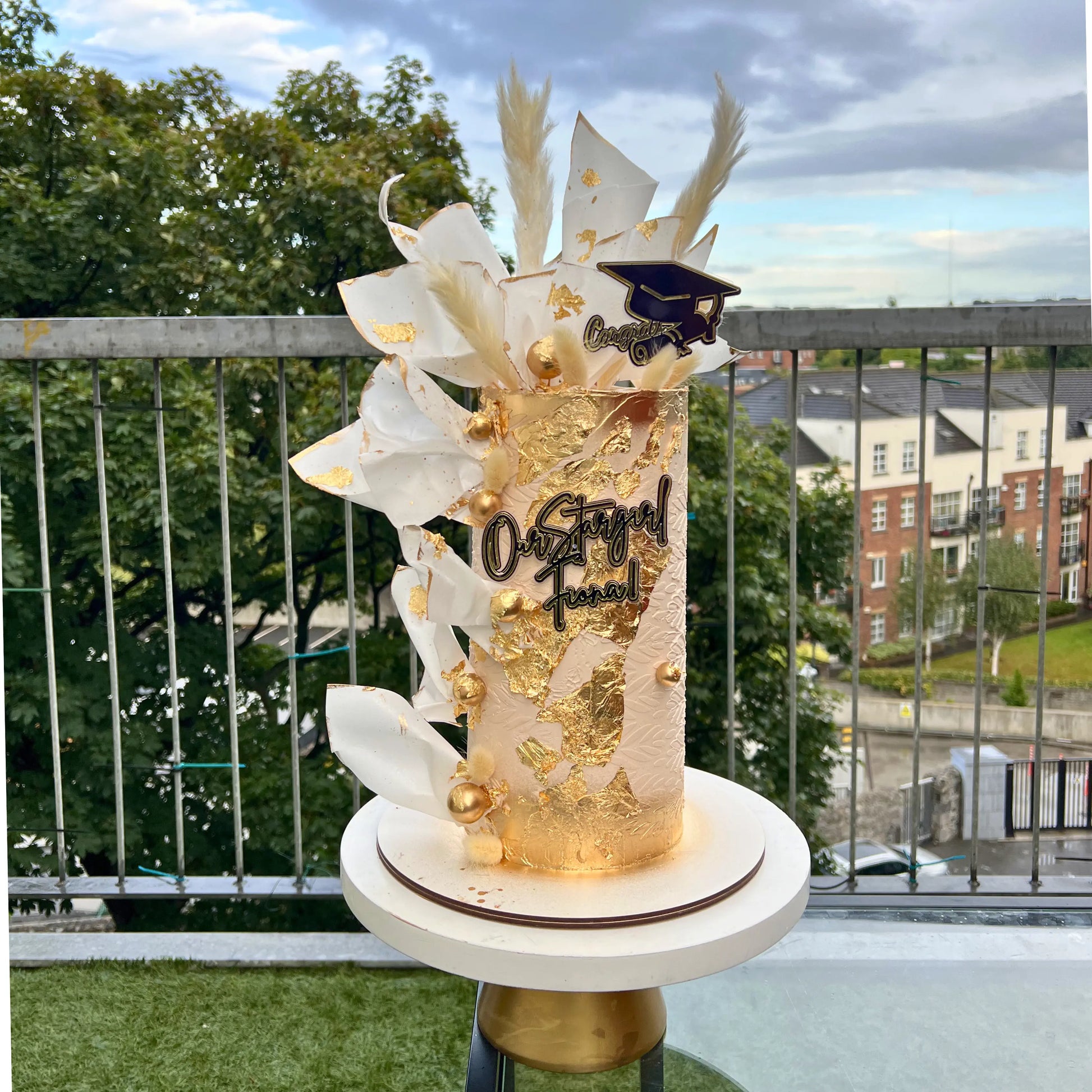 graduation cake with gold accents and 'On Stage' text on a small round table outdoors.on cake 
