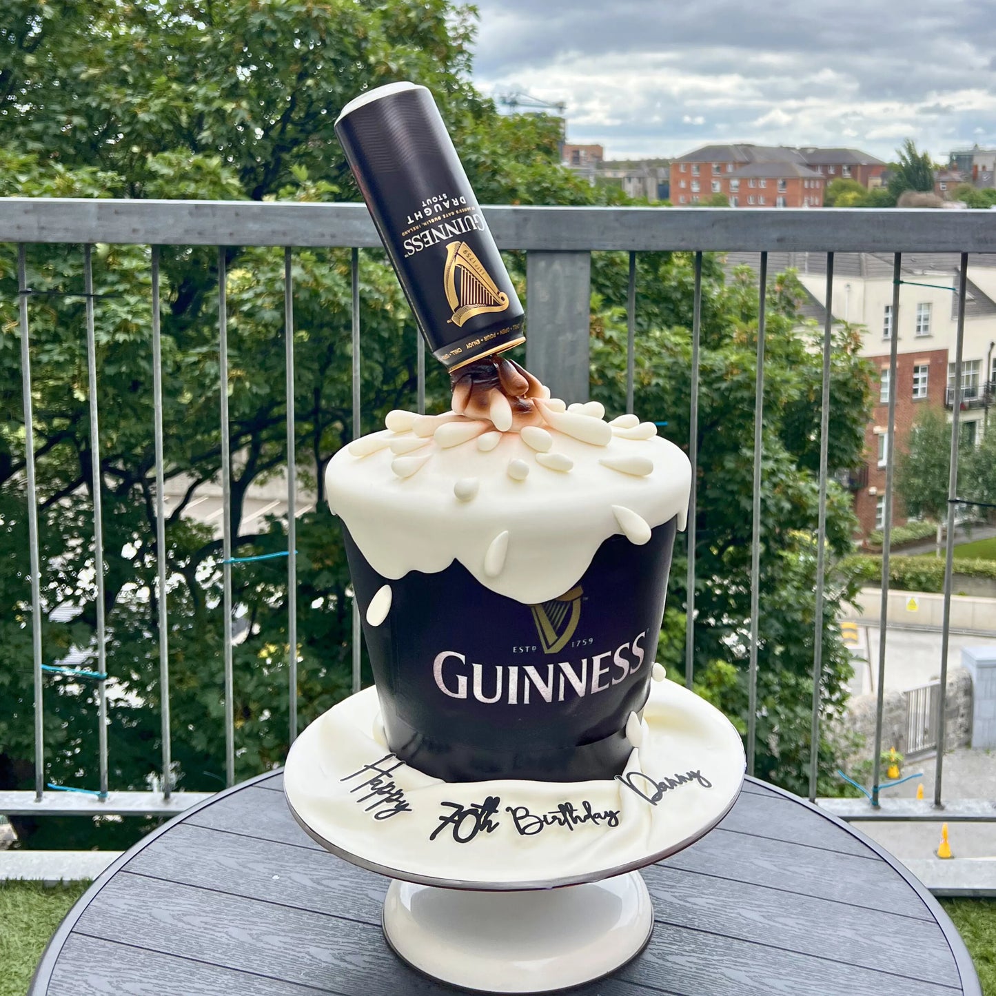 Guinness-themed birthday cake with a bottle on top, outdoors.