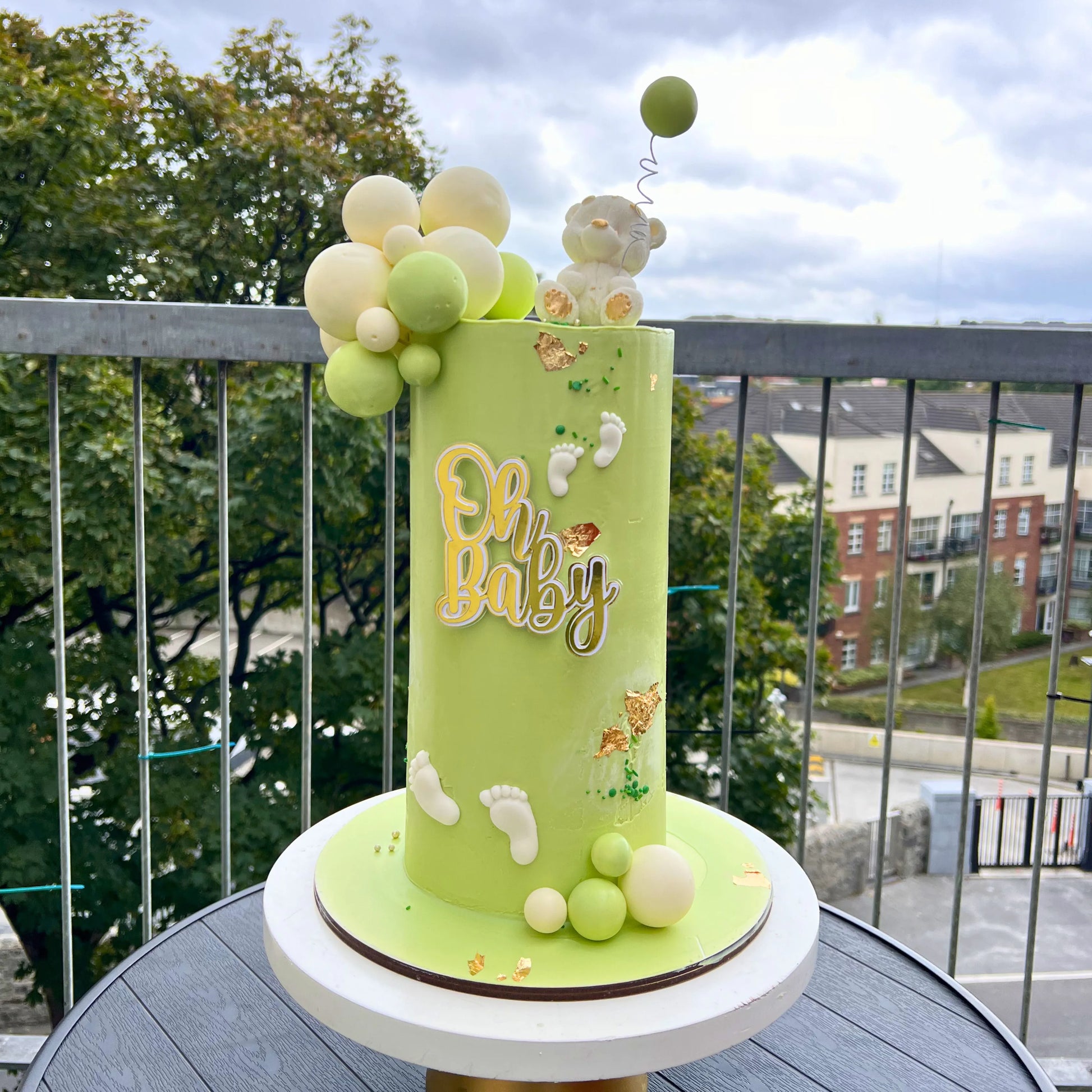 Green baby shower cake with balloons and 'Oh Baby' text on a balcony with trees and buildings in the background.