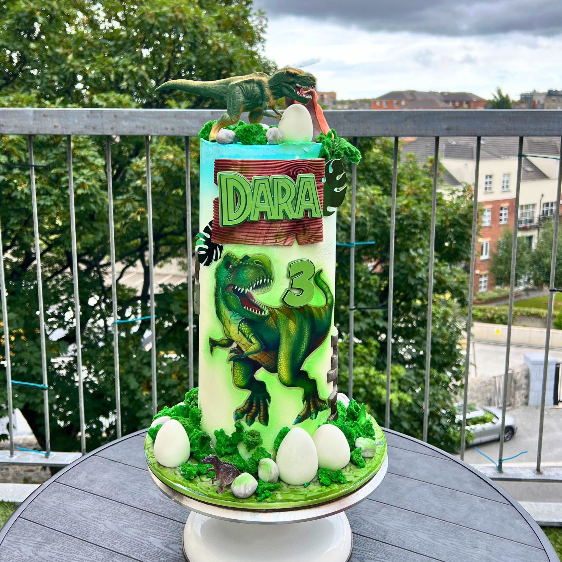 A custom dinosaur-themed birthday cake on a white pedestal stand, set on a balcony with a view of trees and buildings in the background. The cake is decorated with green and white icing, featuring a large green T-Rex, white eggs, a small toy dinosaur, and a wooden-style sign that reads 'DARA'.