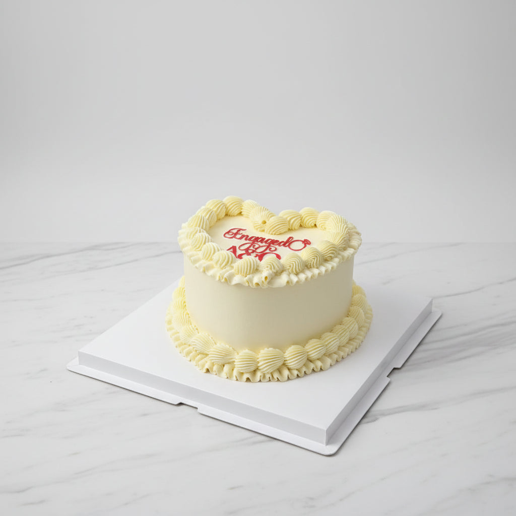 A round white frosted birthday cake with piped cream borders and red icing text reading 'Engaged 28' on top, displayed on a white square cake box with an outdoor city street and apartment buildings in the background.