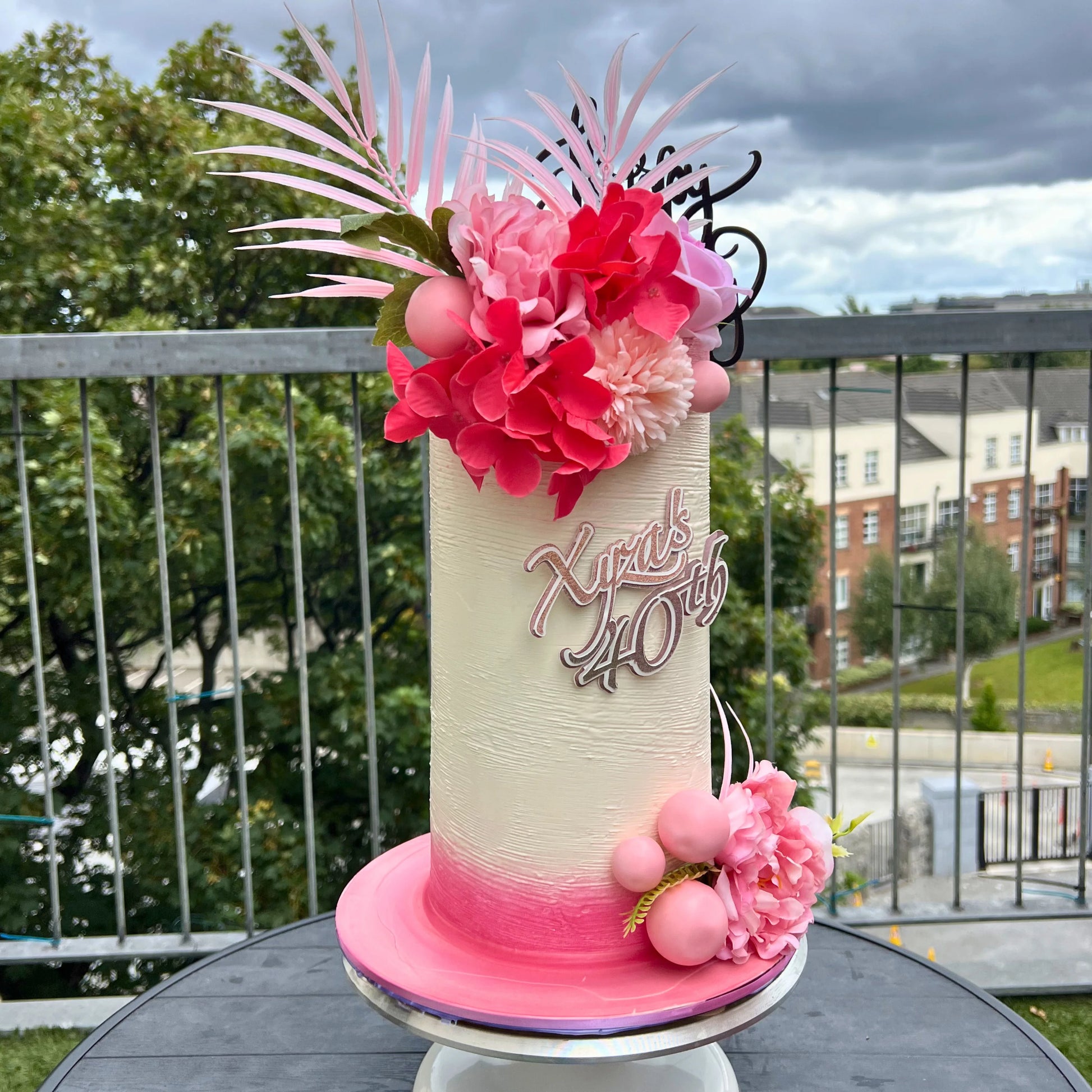 light pink and deep pink birthday cake with floral decorations with pink flowers and 'Happy 40th' text on a table outdoors.