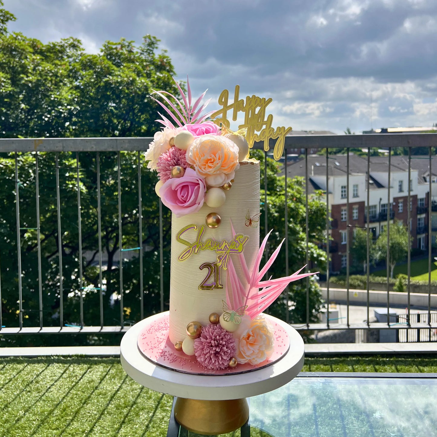 Decorative cake with flowers and 'Happy 21st' topper on a balcony with greenery and buildings in the background