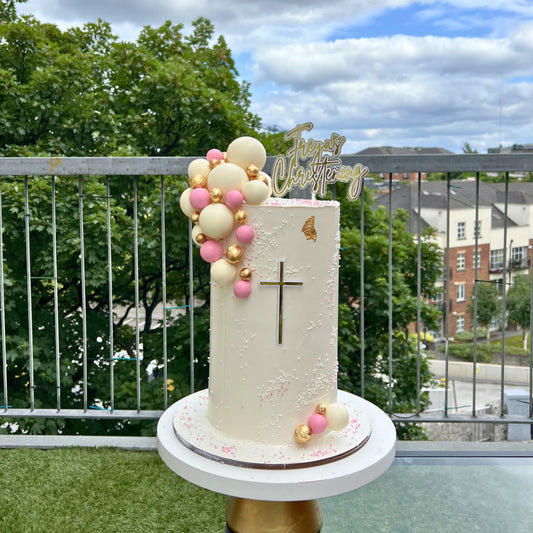 Decorative christening cake with balloons and a cross on a stand outdoors.