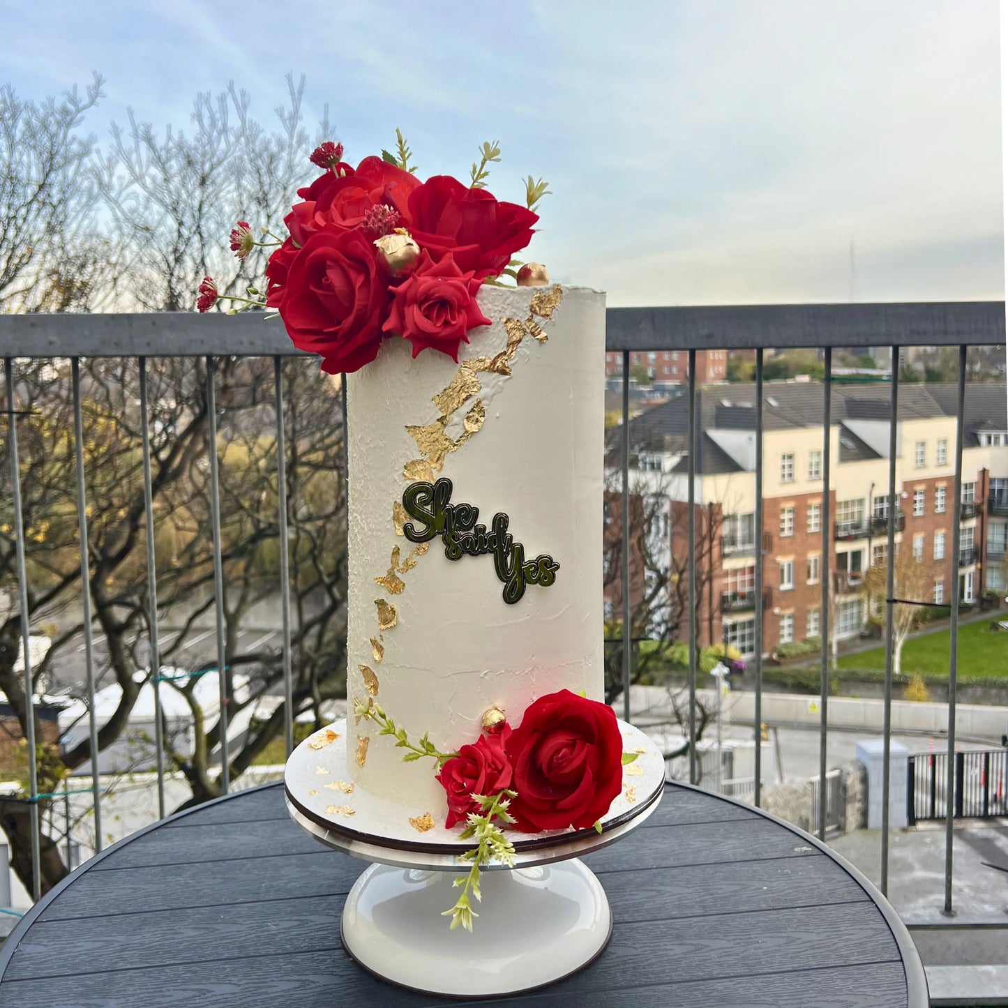 Decorative cake with red flowers and gold accents on a balcony with a cityscape background