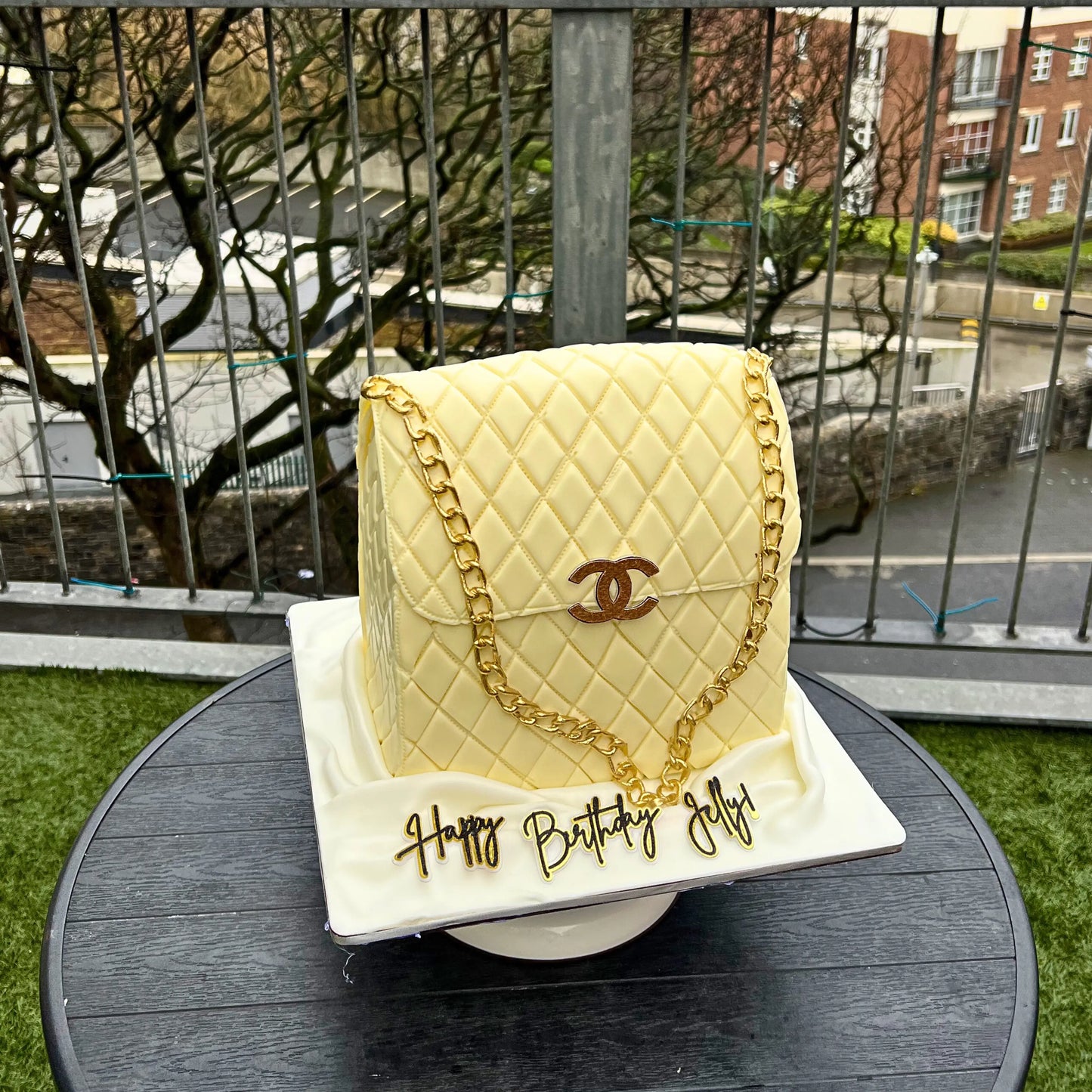 Cake designed like a quilted handbag with gold chain and 'Happy Birthday Jelly' text, outdoors.