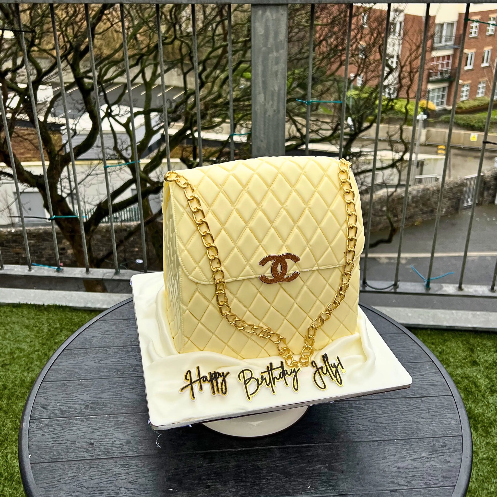 Cake designed like a quilted handbag with gold chain and 'Happy Birthday Jelly' text, outdoors.