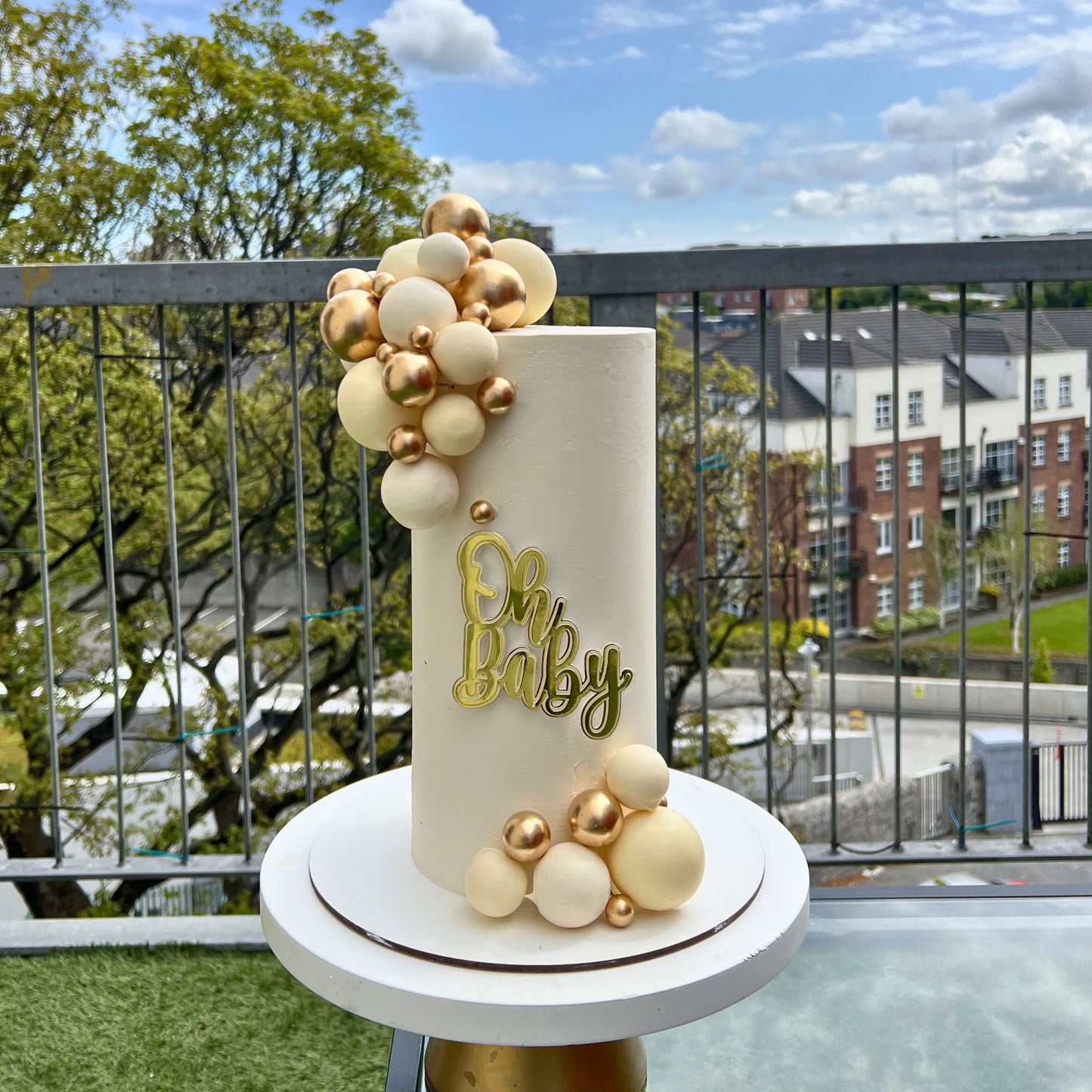Decorative cake with 'Oh Baby' text on a balcony with a view of buildings and trees.