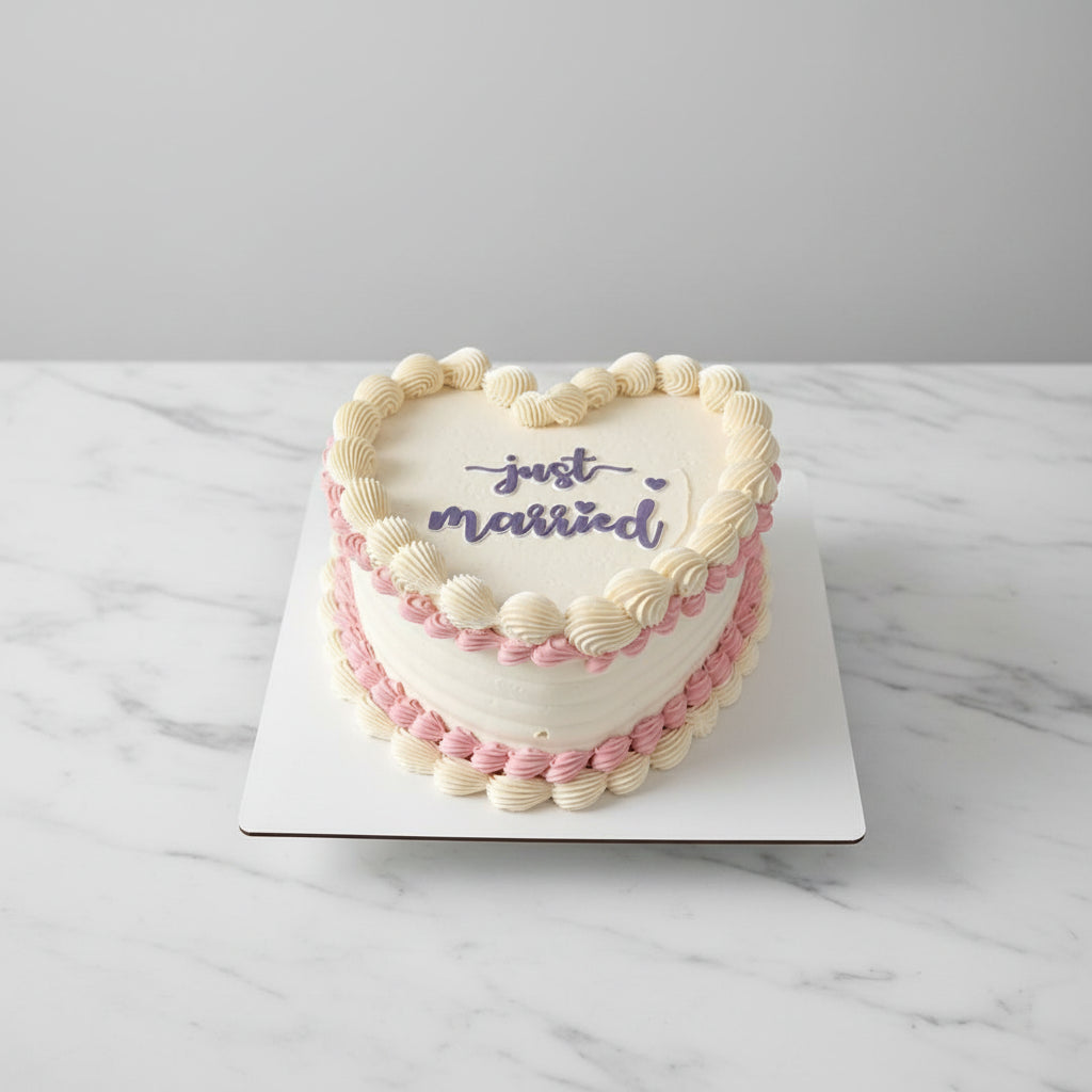 A heart-shaped frosted cake on a white board, decorated with cream and pink piped borders and the words 'just married' in purple script, photographed from above over an outdoor residential street scene.