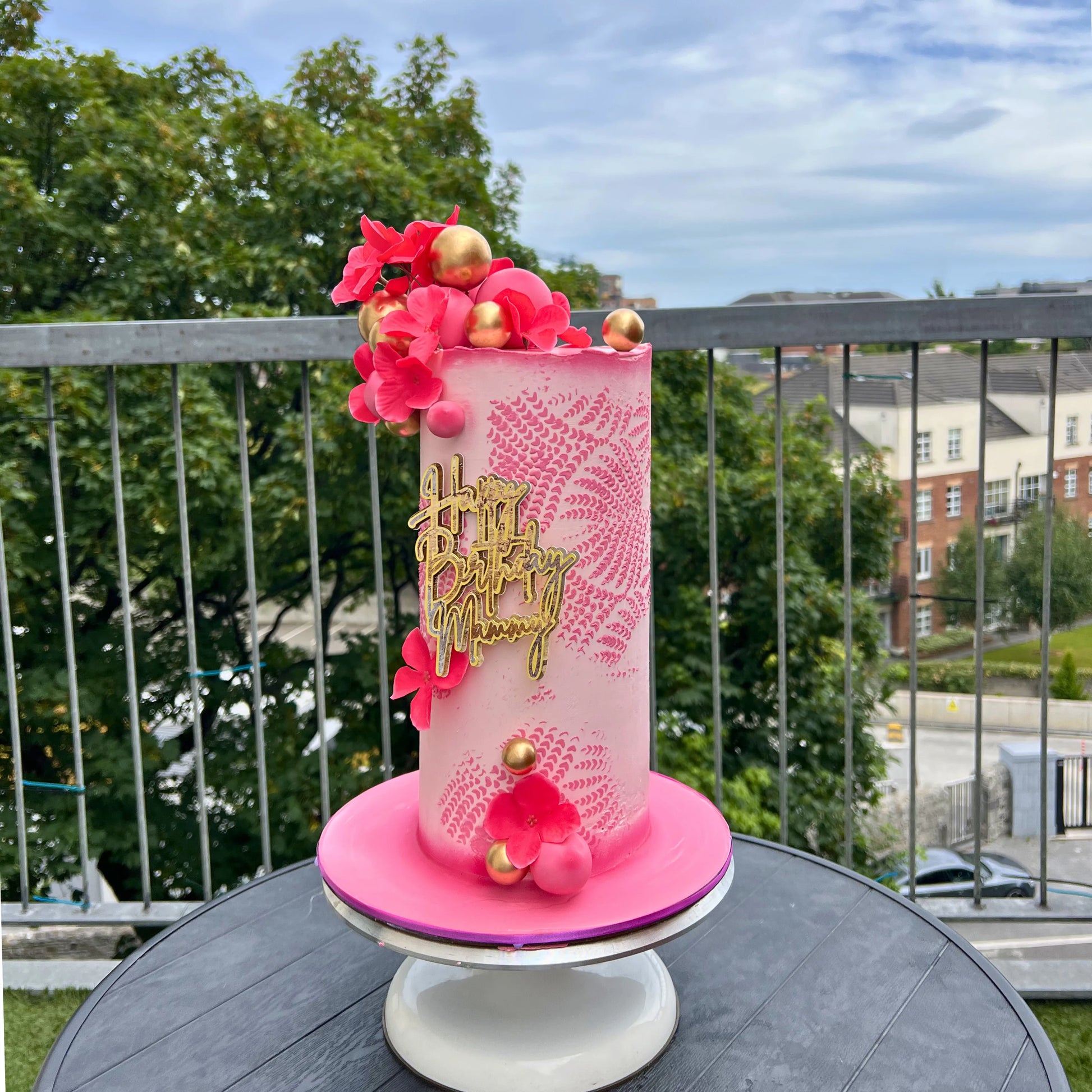 light pink and deep pink birthday cake with floral decorations with pink and gold design on a stand outdoors.