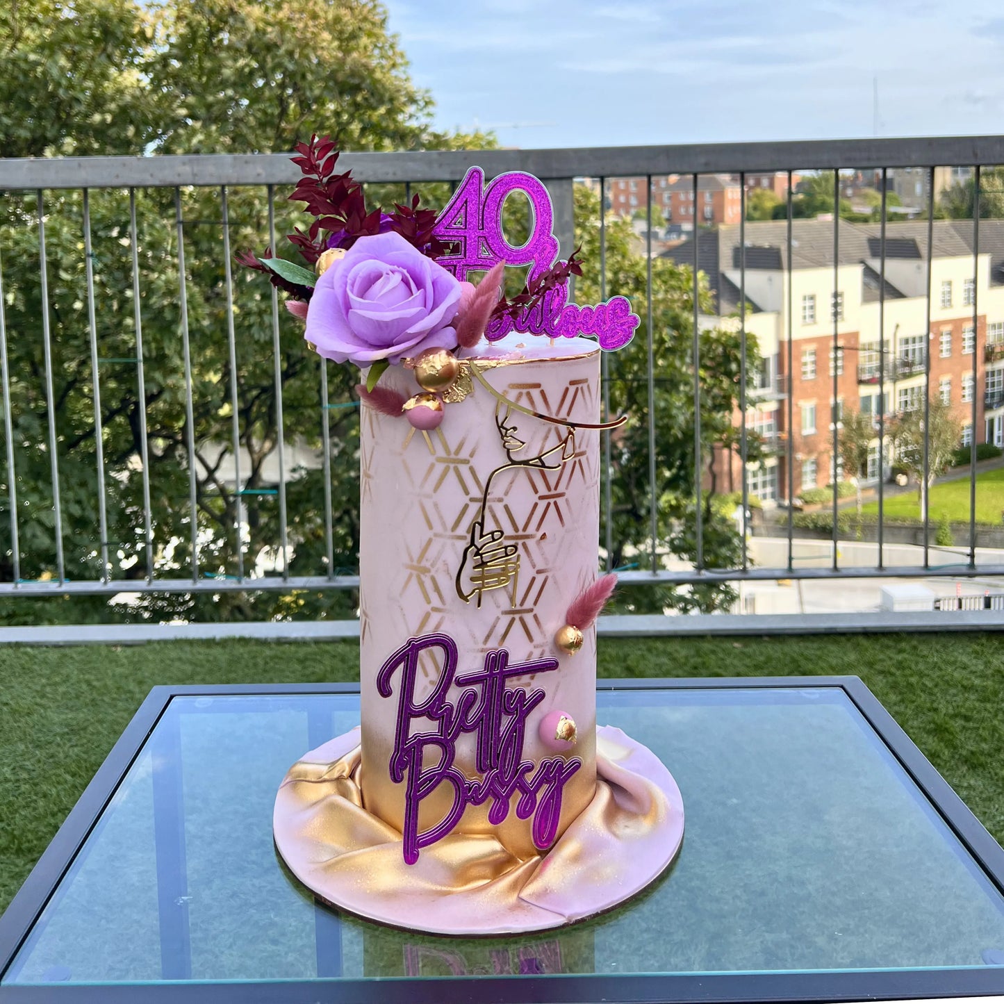lilac  birthday cake with floral decorations with '40' and 'Betch Betch' text on a table outdoors.