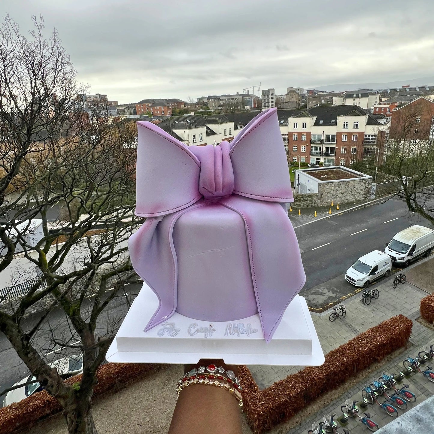 Purple cake with a large bow on top, held up against an urban landscape.