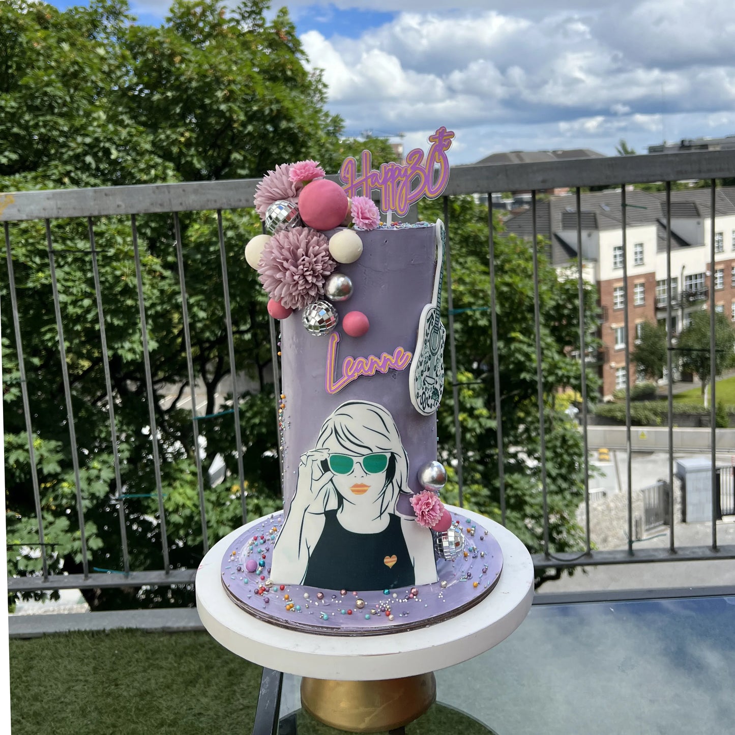 Decorative cake with a portrait and floral elements on a balcony with greenery and buildings in the background.