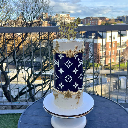 Designer cake with gold accents and 'Happy Birthday' topper on a balcony with cityscape background
