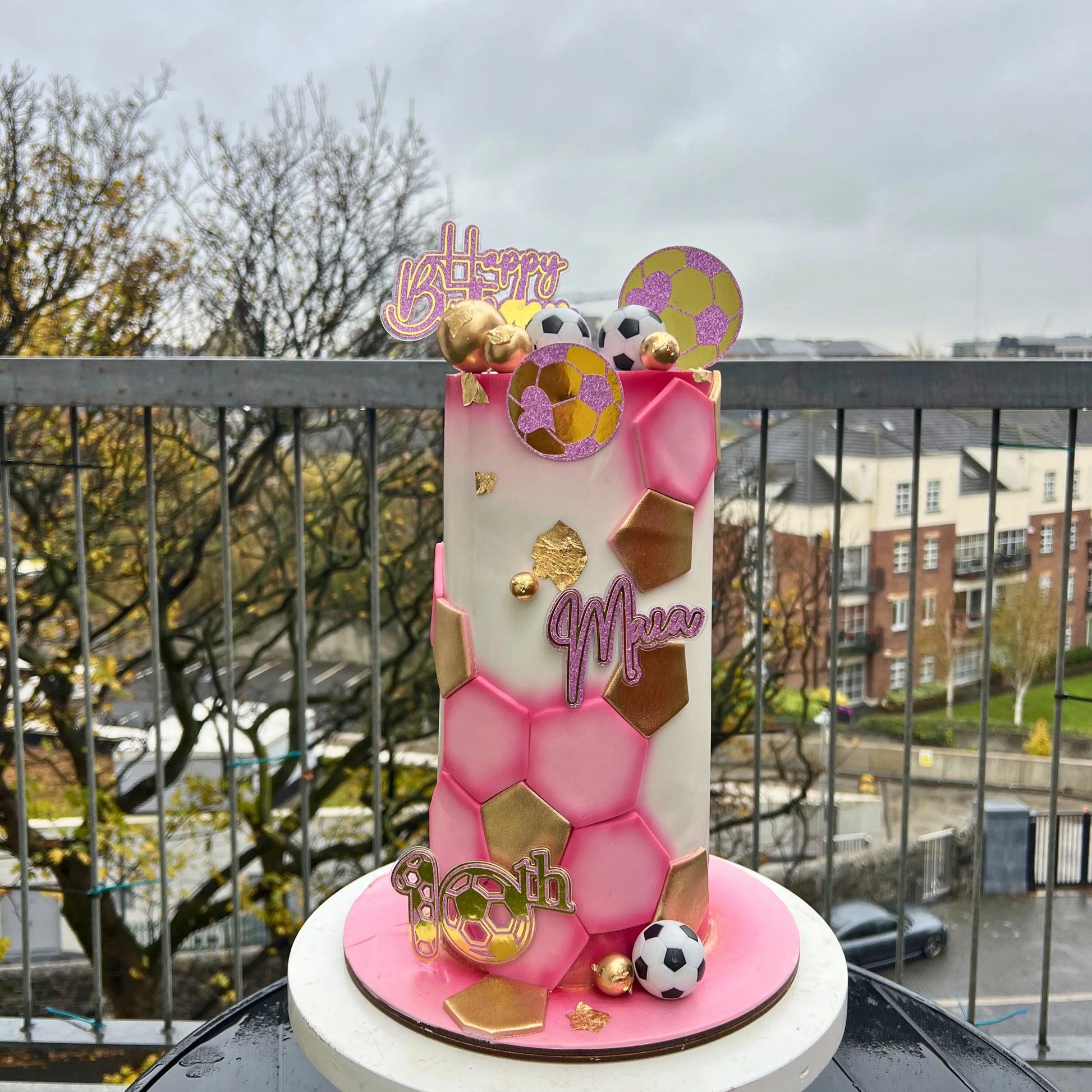 Decorative cake with pink and gold design on a balcony with a view of buildings and trees.