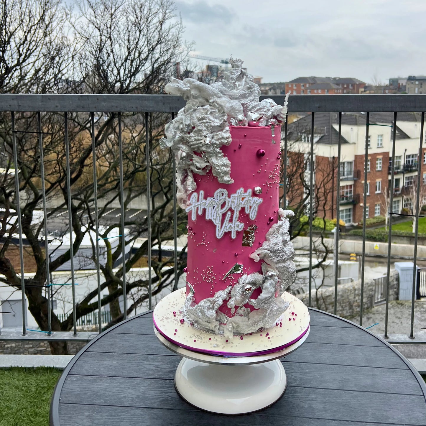 Pink birthday cake with silver decorations on a table outdoors.