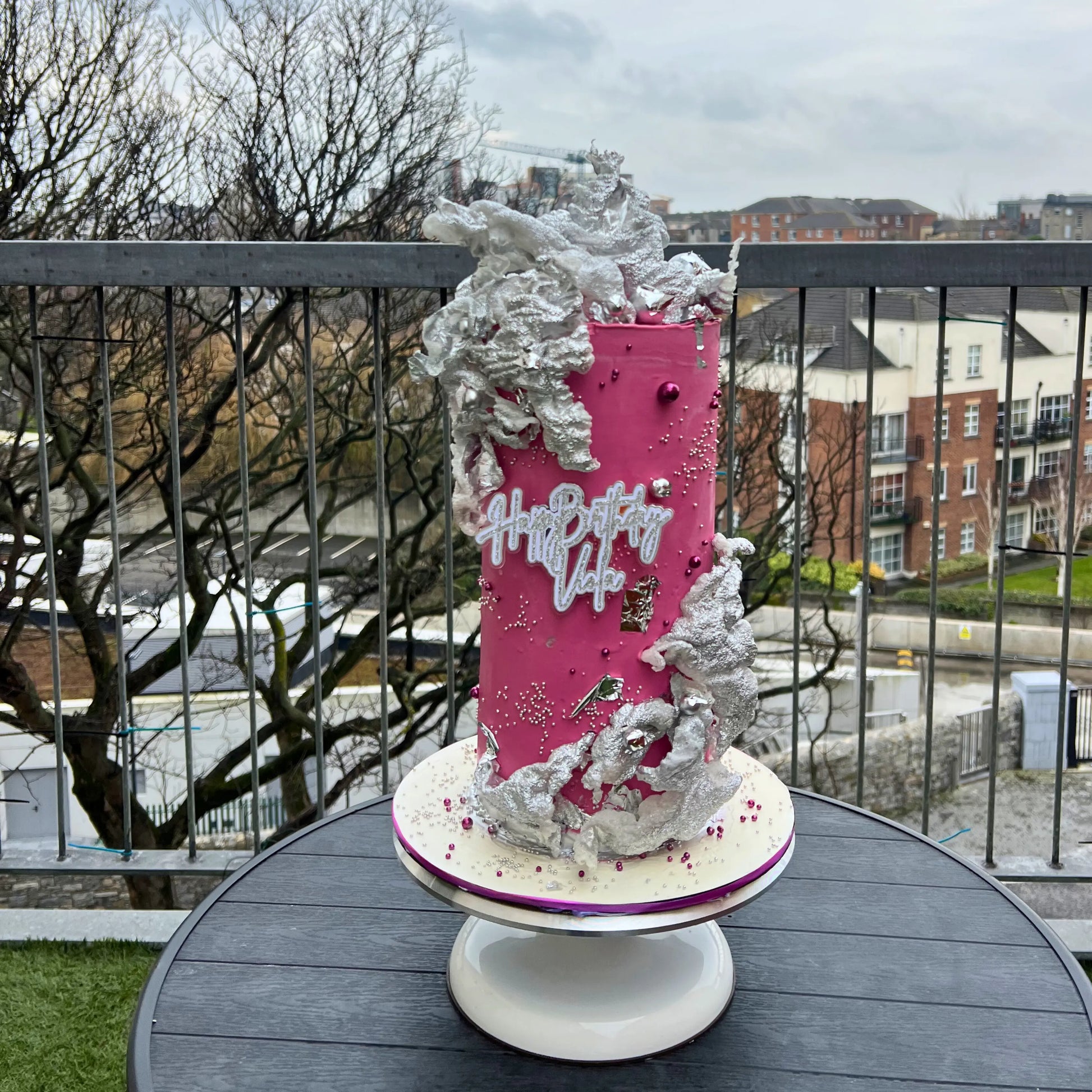 Pink birthday cake with silver decorations on a table outdoors.