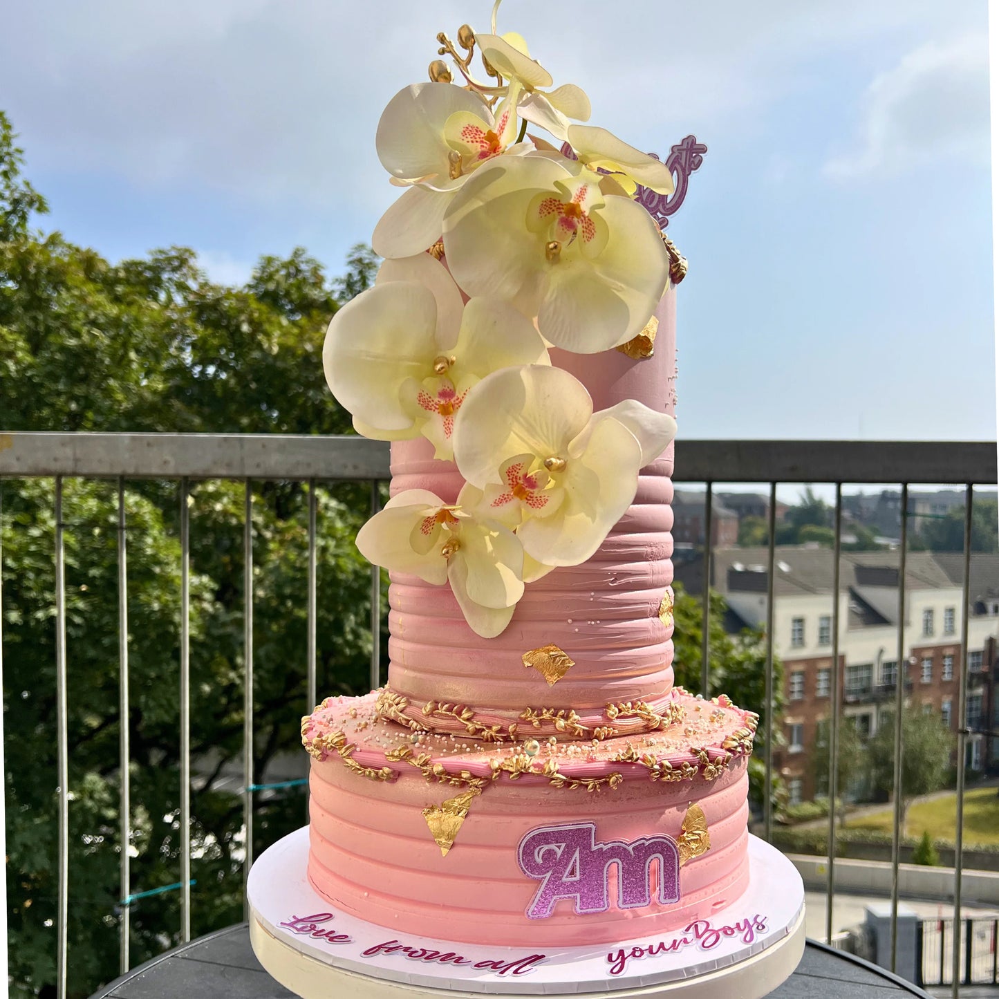 Pink cake with yellow flowers on a balcony with a view of trees and buildings.
