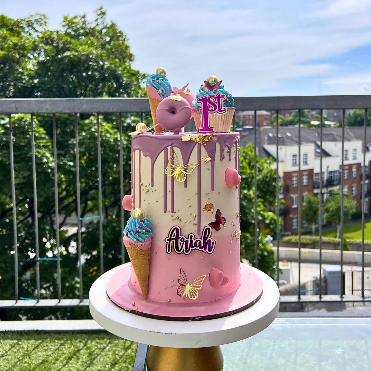 Pink birthday cake with ice cream cones and 'Ariah' on a balcony with greenery and buildings in the background