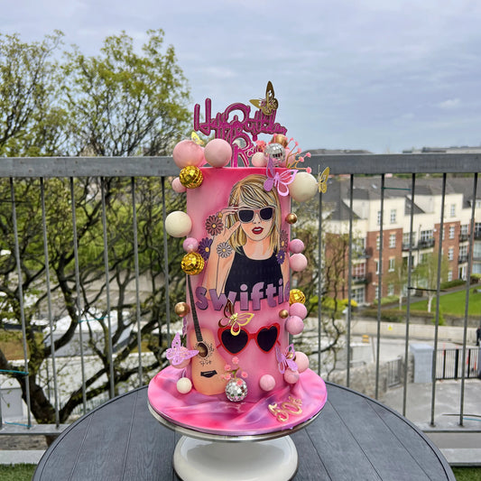 Pink taylor swift birthday cake with decorative elements on a table outdoors.