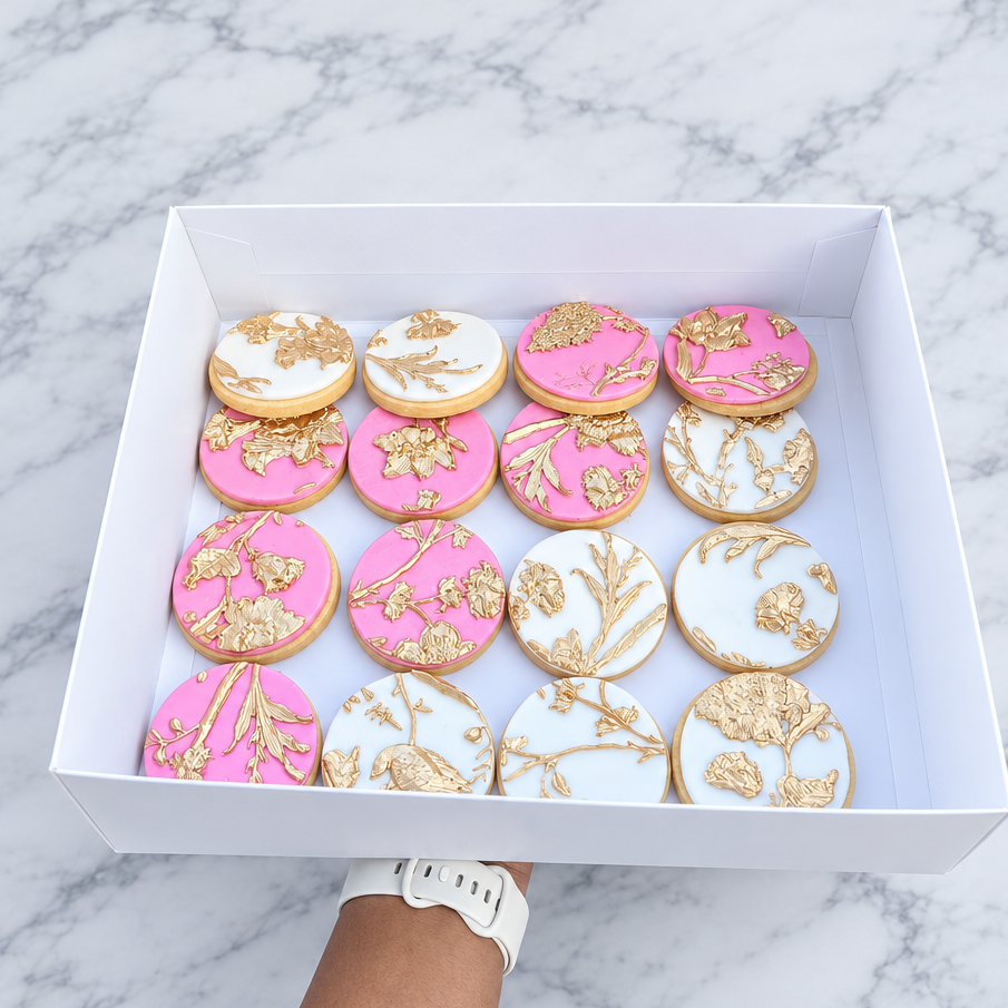 A white rectangular box containing 15 round iced cookies with pink, white, and gold floral designs, held up by a person's arm outdoors over a garden and driveway.