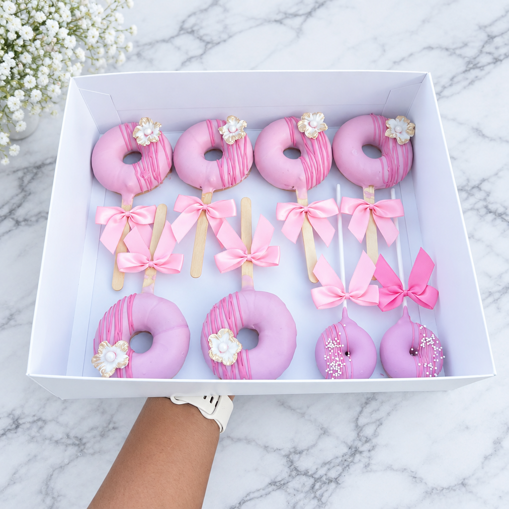 A white gift box filled with pink donut-shaped treats and pink ribbon lollipops, held by a person's hand outdoors over a balcony railing.