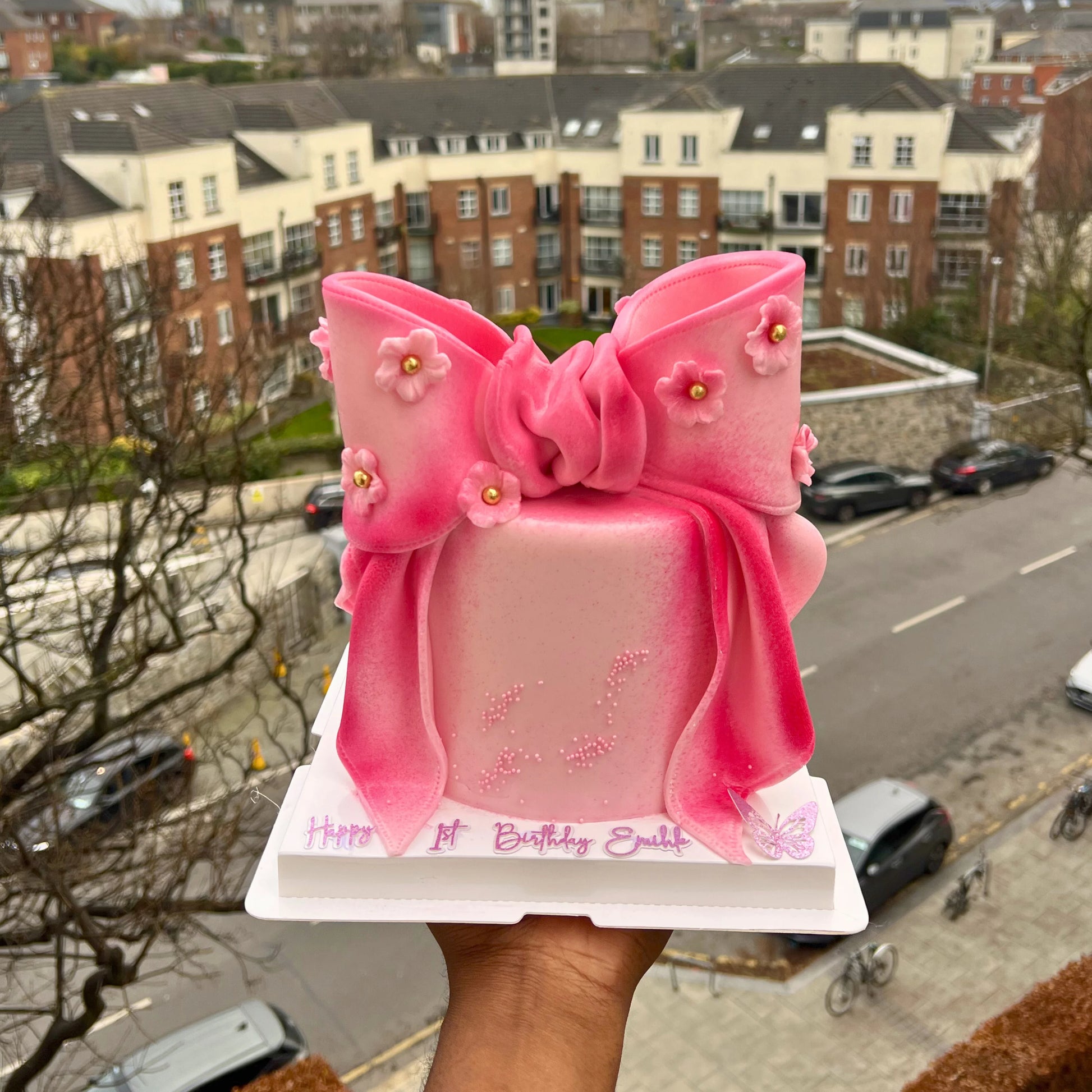 Pink birthday cake with a large bow held up against an urban background