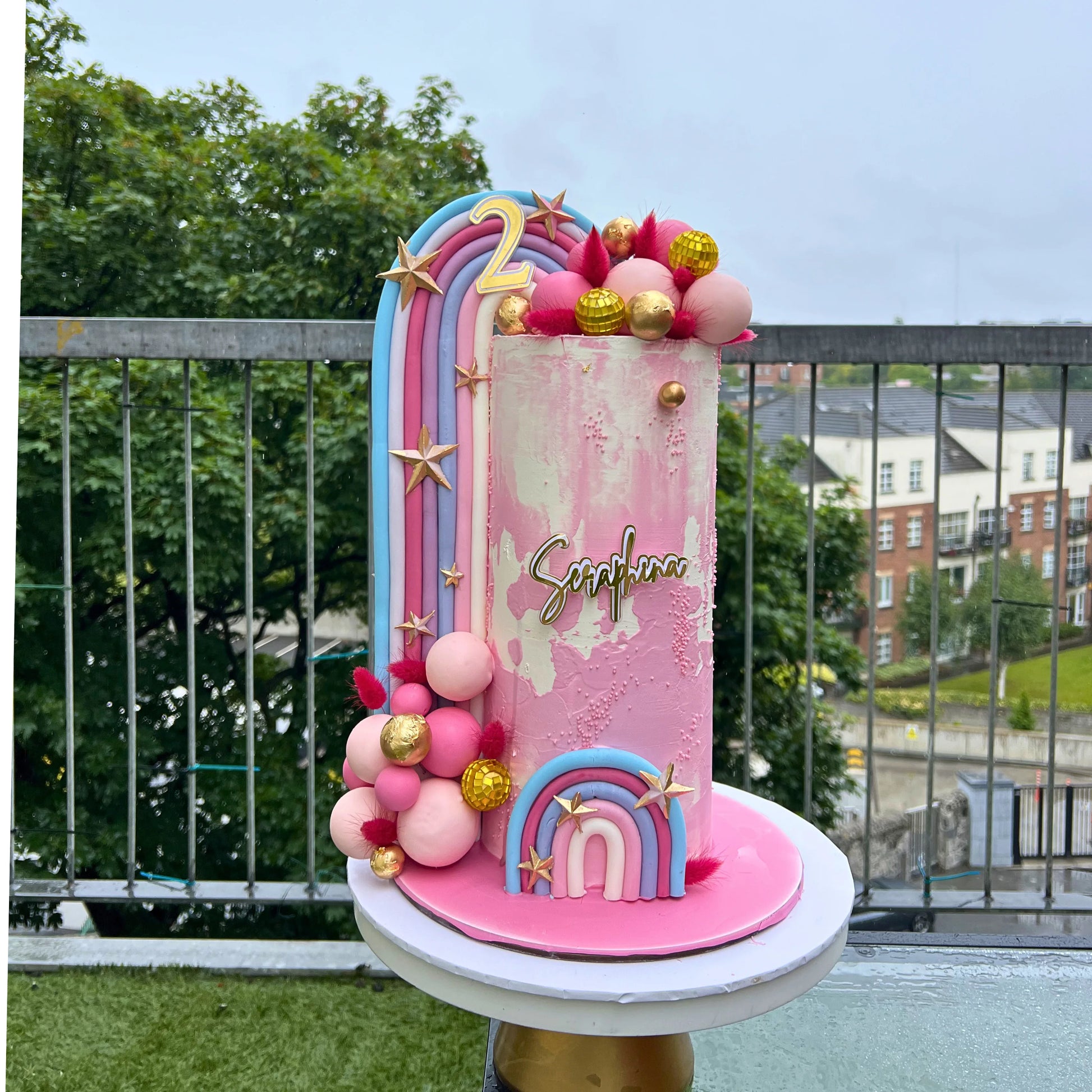 Colorful birthday cake with rainbow and star decorations on a rooftop with a cityscape background.