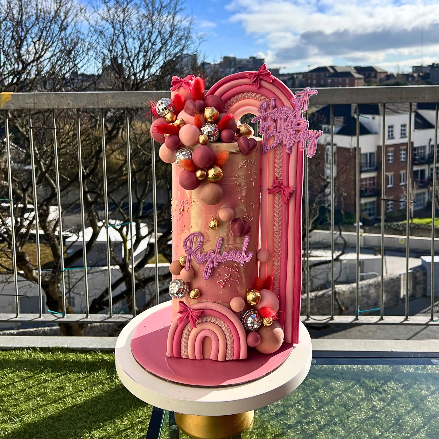 Decorative pink cake with colorful embellishments on a stand outdoors.