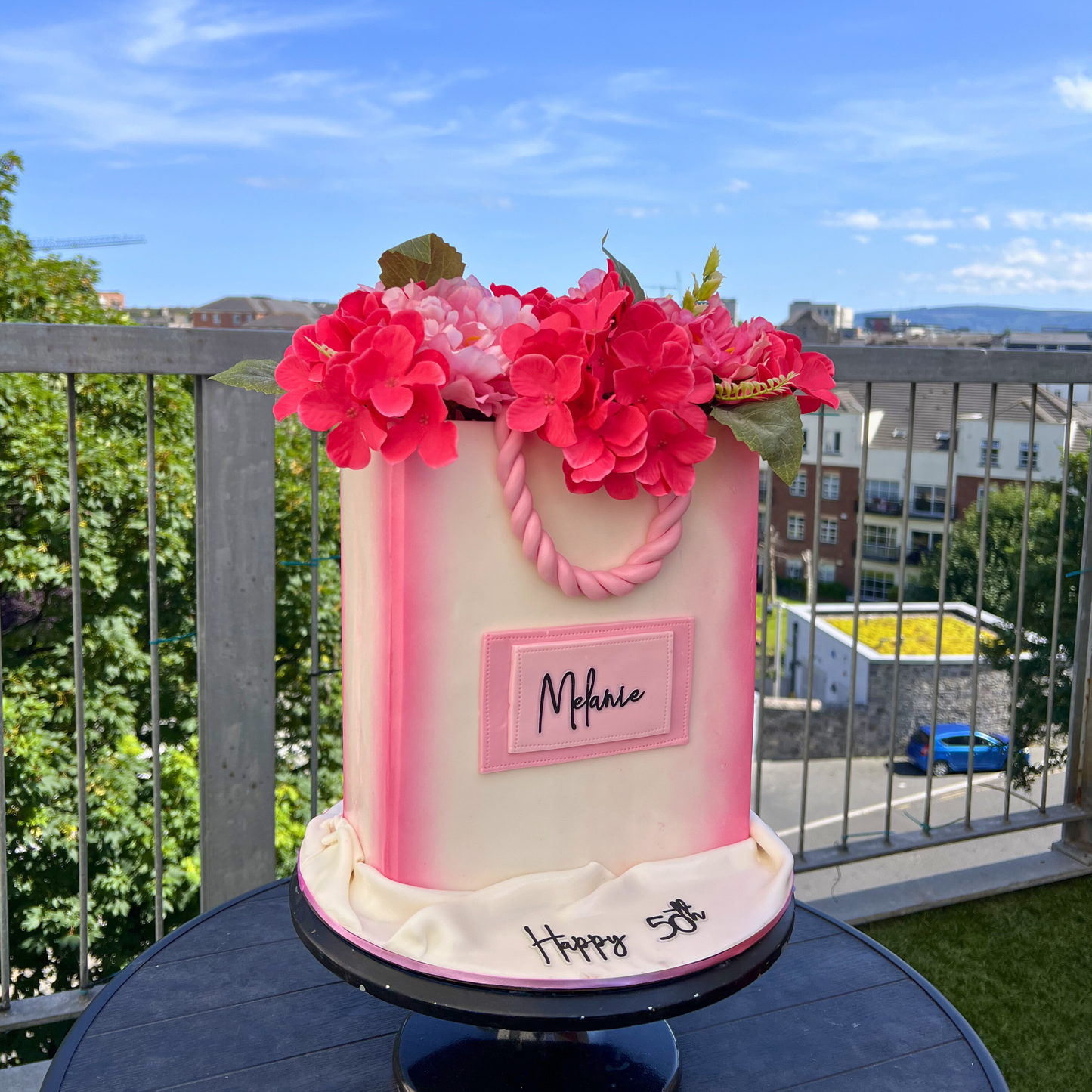 Decorative cake with pink flowers and 'Melanie' on a balcony with a cityscape background
