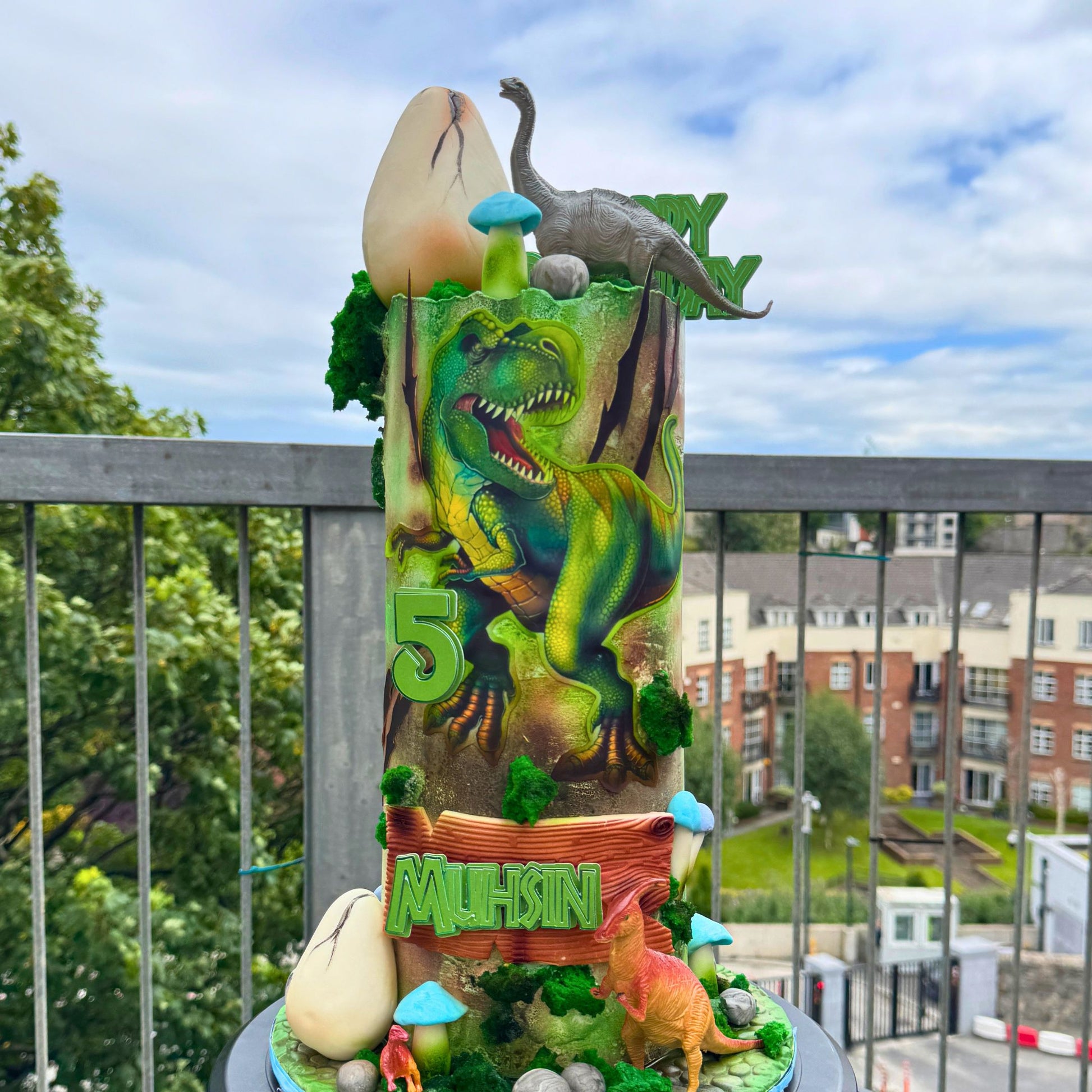 A tall dinosaur-themed birthday cake with a green T-rex, a large cream-colored egg, blue mushrooms, green lettering, and a small orange dinosaur decoration