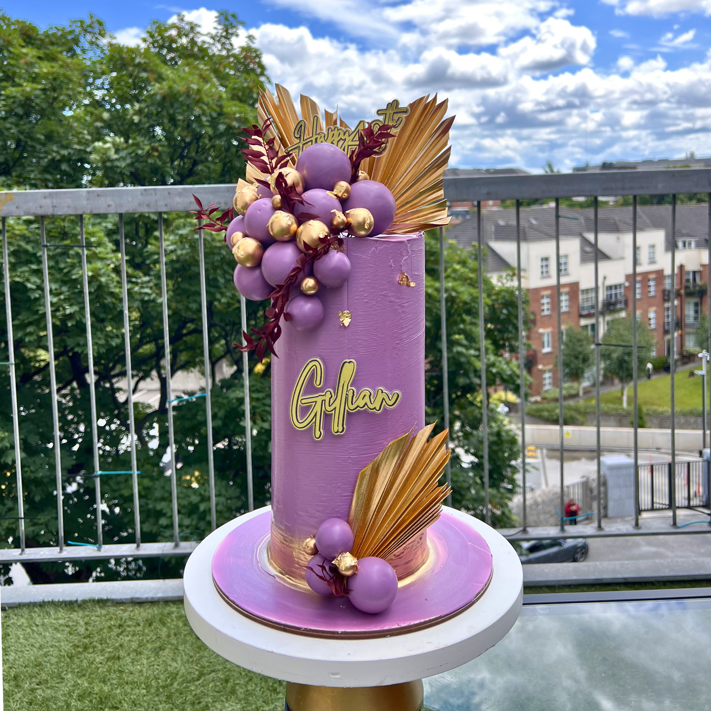 Decorative cake with purple and gold elements on a balcony with a view of buildings and trees.