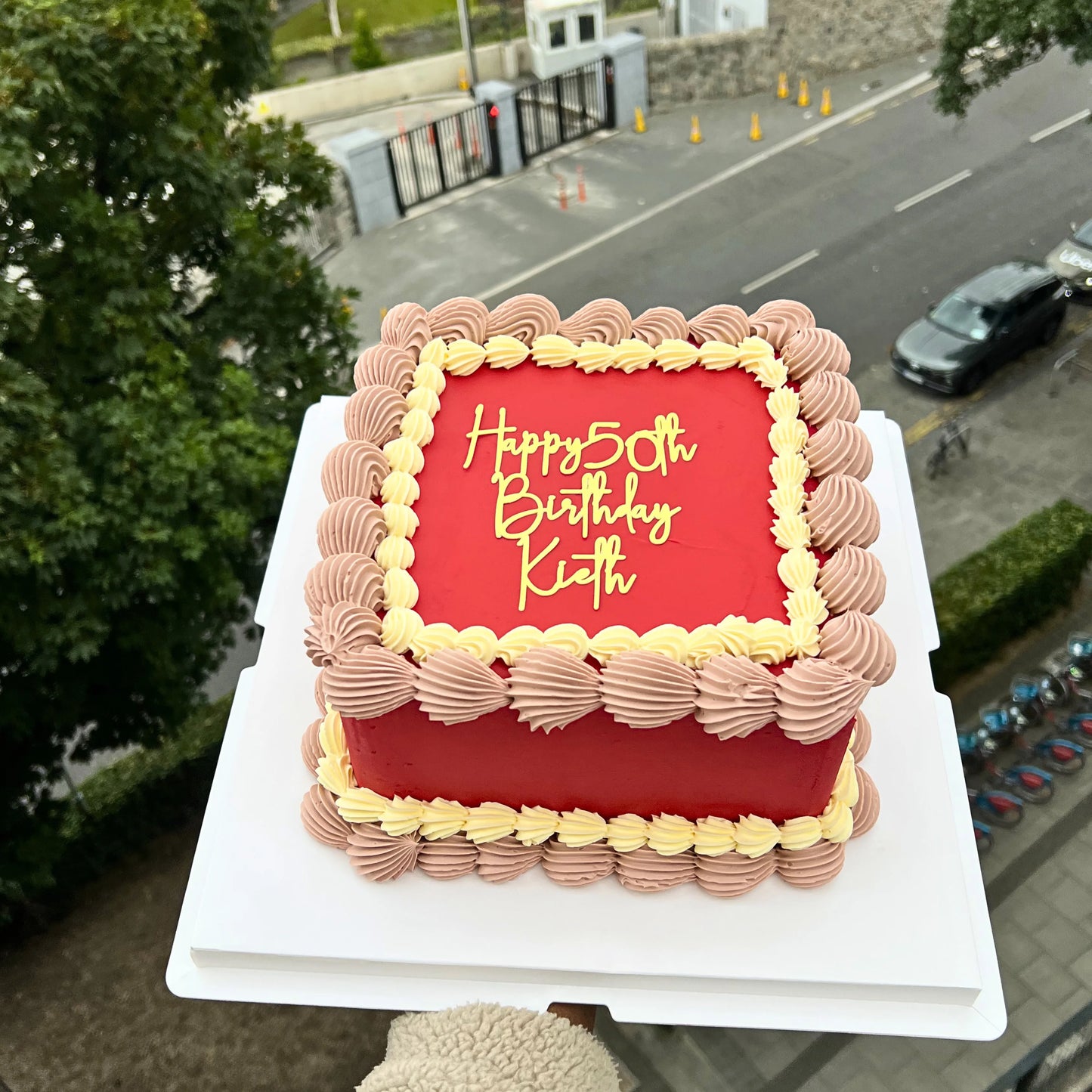 Red birthday cake with gold lettering on a white stand, outdoors with road and trees in the background