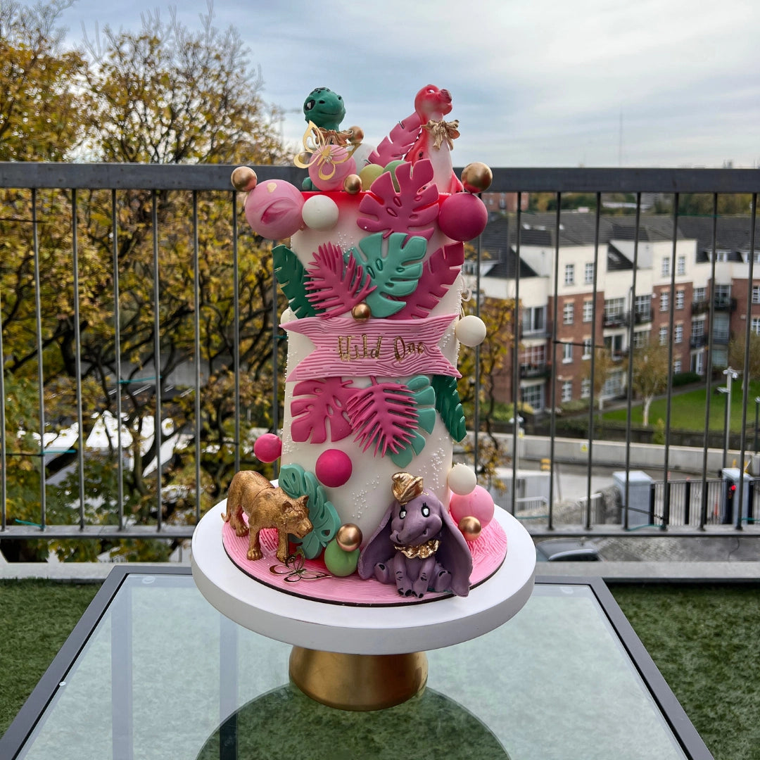 A decorated cake with a safari theme, featuring animal figurines and tropical leaves, displayed on a table outdoors.