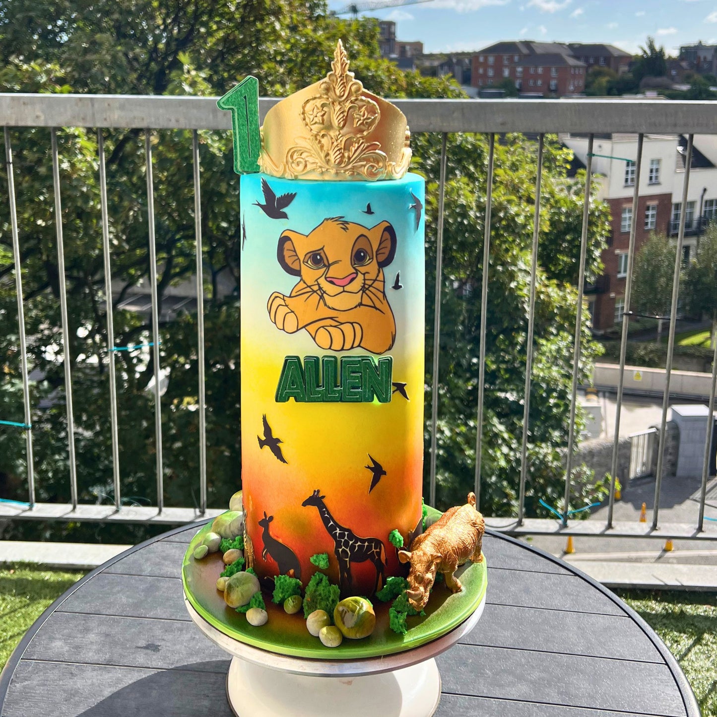 A custom birthday cake shaped like a tall cylinder, decorated with a Simba cub illustration, the name 'ALLEN' in green letters, and safari-themed details including a gold crown on top, a green base with animal silhouettes, and a small lion figurine, displayed on a balcony table.