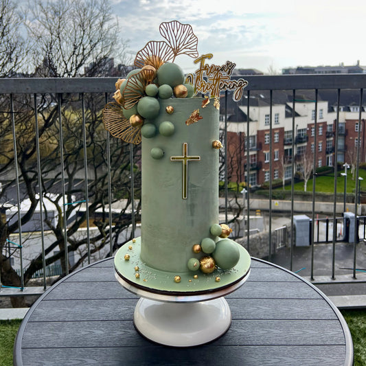 sage christening cake with green fondant, gold accents, and a cross on a stand outdoors.